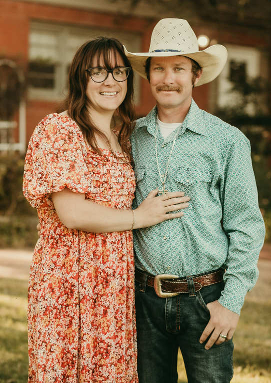 A smiling couple standing outdoors in front of a house, with the woman wearing glasses and a floral dress and the man wearing a cowboy hat, a patterned button-up shirt, and a belt with a large buckle.