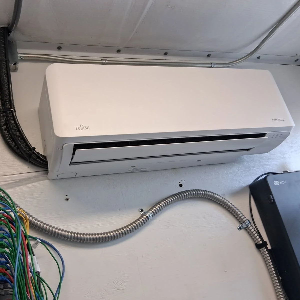 White Fujitsu wall-mounted air conditioning unit installed on a white wall with visible electrical wiring and conduit pipes.