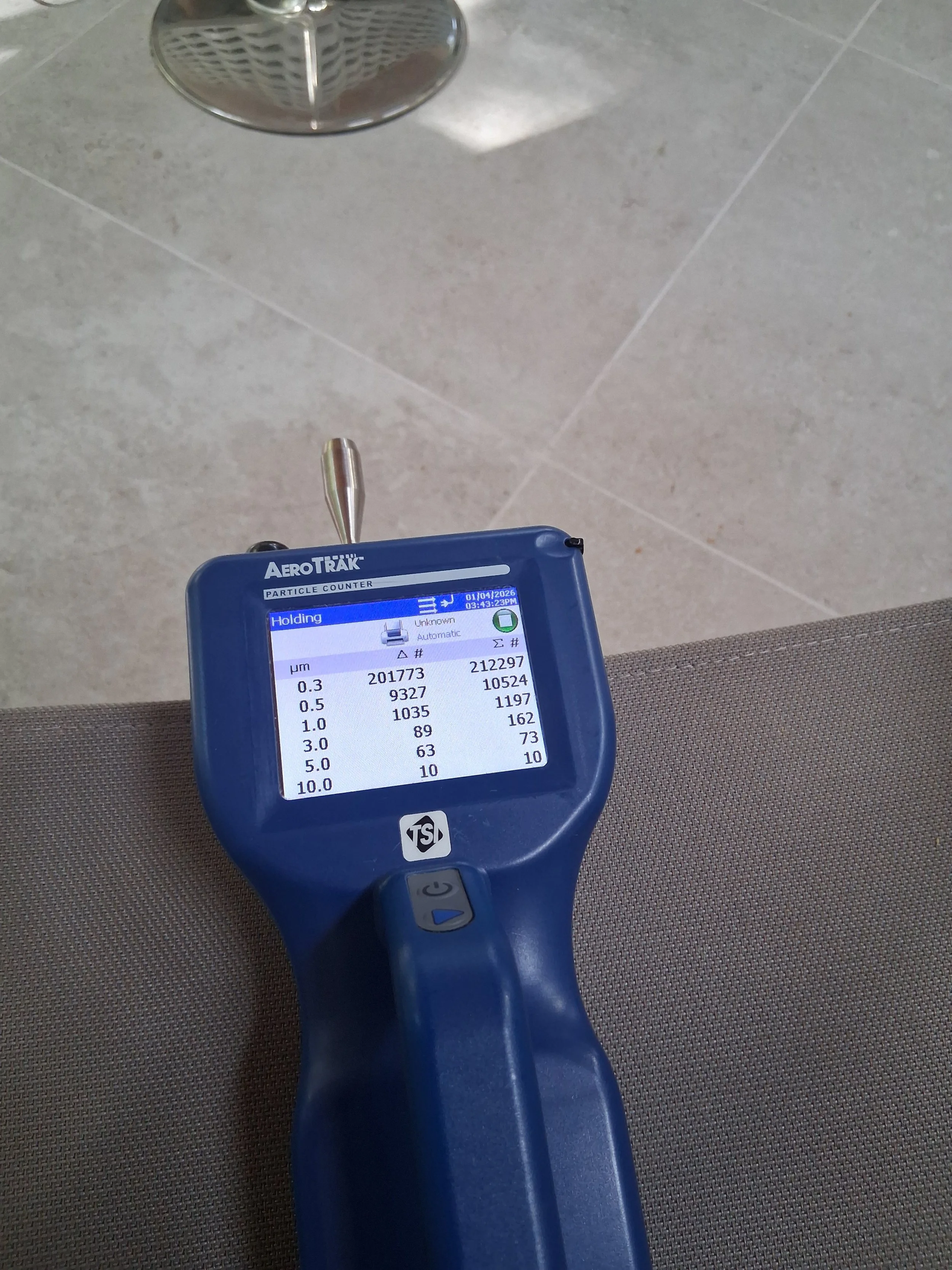 A handheld particle counter device with a blue body and a small display screen showing numerical data, held above a gray surface with a metallic drain in the background.