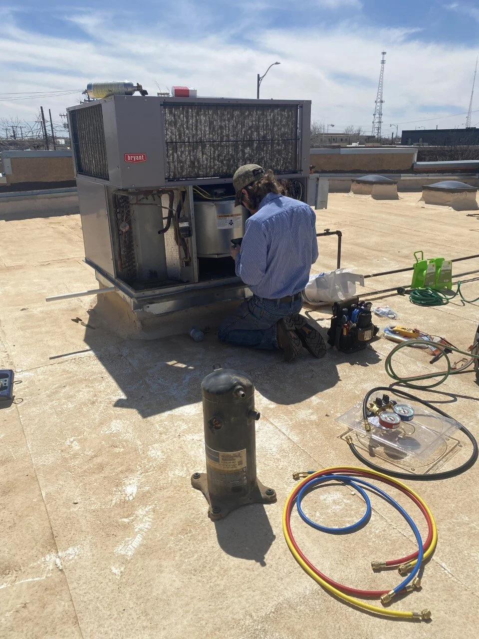 A technician kneels on a rooftop servicing an HVAC unit with various tools and refrigerant hoses nearby, under a partly cloudy sky.