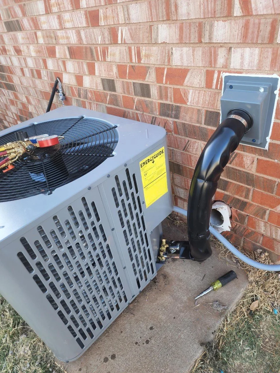 Outdoor HVAC unit installed outside a brick wall, with black insulated piping connected to it, a screwdriver on the concrete, and some dried grass nearby.