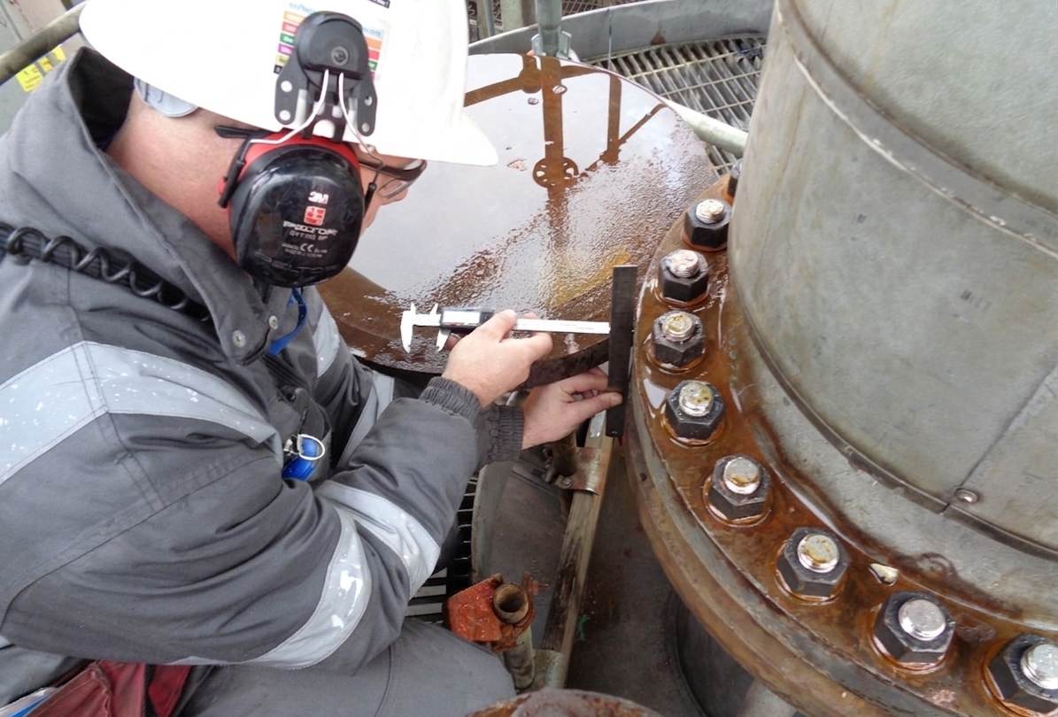 Worker in safety gear, including hard hat, ear protection, and gloves, using a caliper to measure a large industrial metal pipe with bolts.