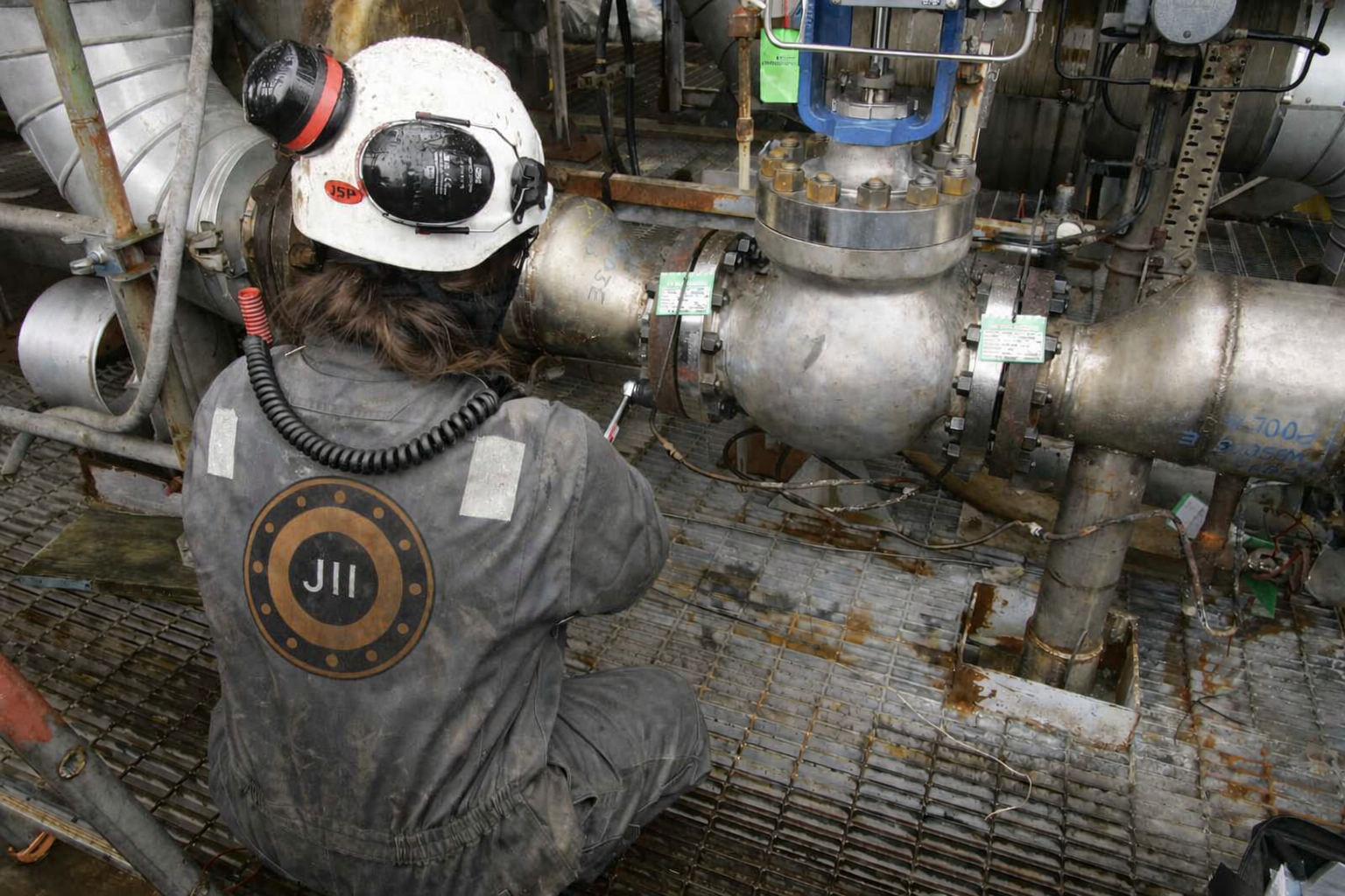 A worker wearing a helmet and gray jumpsuit with JII logo is working on industrial piping, possibly in a factory or facility. The worker is using tools to inspect or repair the large metal pipe system.