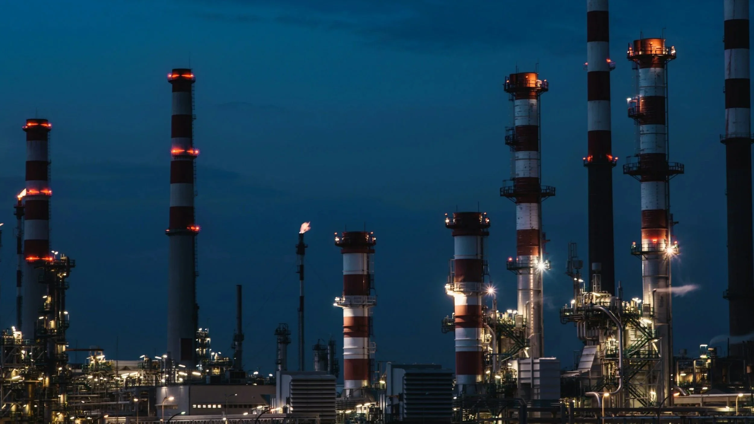 Nighttime view of an industrial oil refinery with multiple tall smokestacks emitting smoke, lit with various lights and orange safety markings.