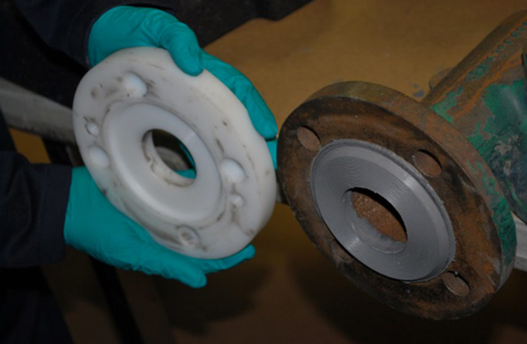 Person wearing blue gloves holding a white ceramic or plastic gasket or seal near a rusty metal pipe or machinery part.