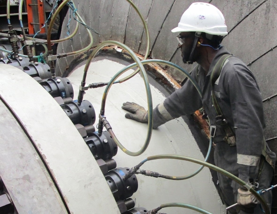 A construction worker wearing a white hard hat, safety goggles, gloves, and gray protective clothing working on industrial equipment with multiple hoses and connectors.