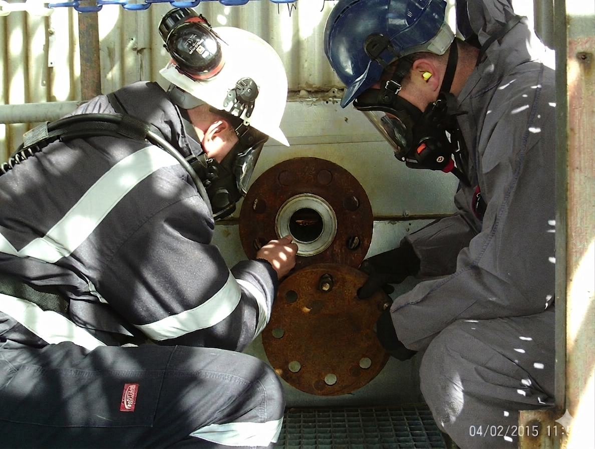 Two workers in protective gear inspecting or working on rusted steel pipe flanges in an industrial setting.