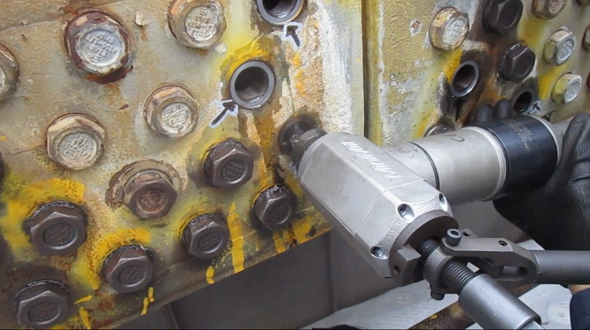 A worker is using a hydraulic bolt removal tool to remove bolts from a rusty, yellow-painted metal surface with multiple bolts.