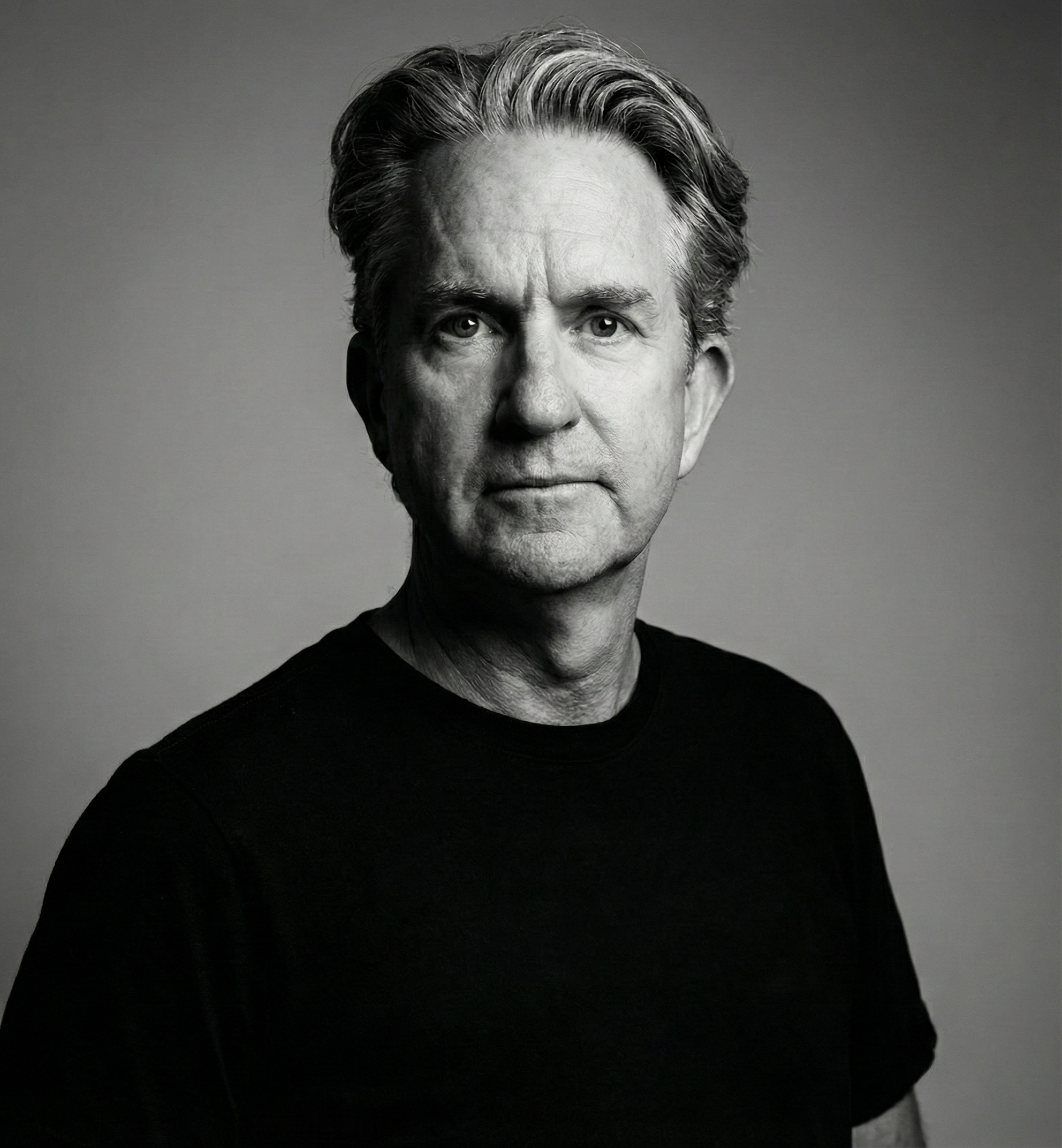 Black and white portrait of a middle-aged man with short, wavy hair. He is wearing a black shirt and is looking directly at the camera with a serious expression. The background is plain and neutral.