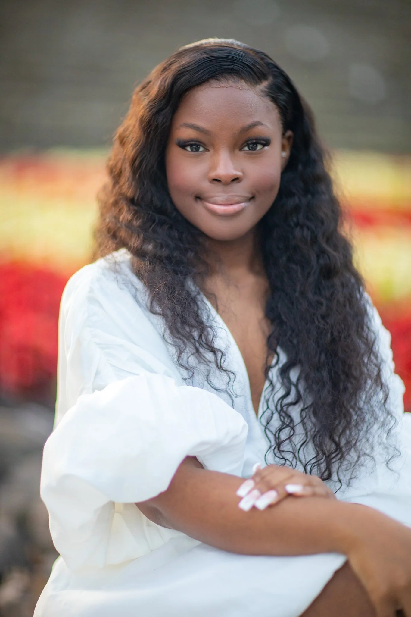 A young African American woman with long, curly black hair, wearing a white blouse, smiling with arms crossed, outdoors with blurred colorful foliage in the background.
