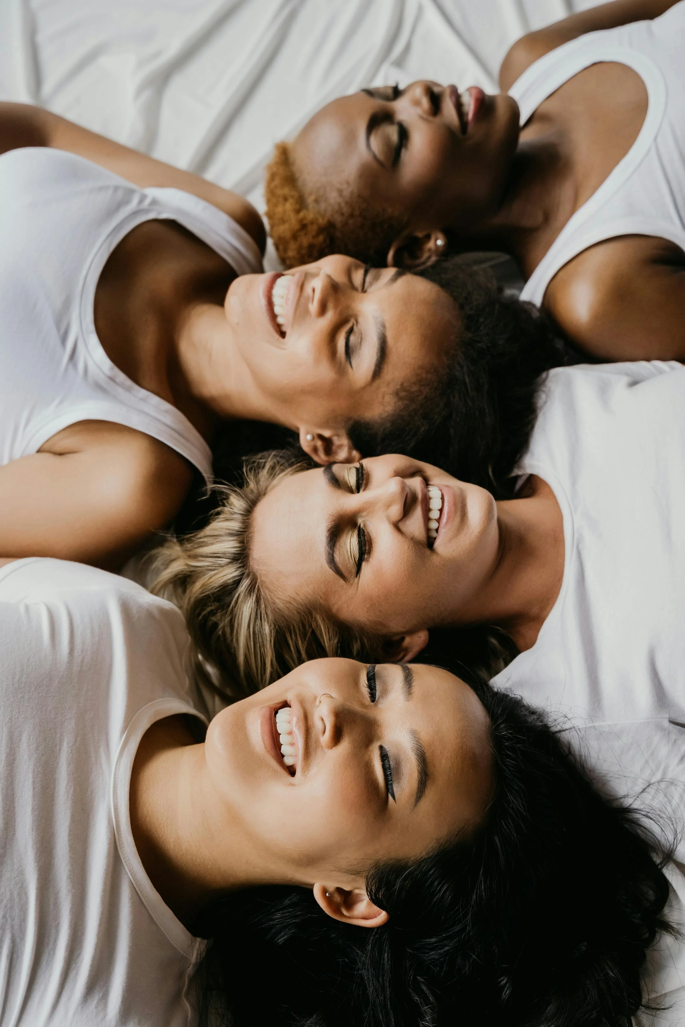 A group of women lying on a white bed, smiling and resting with their eyes closed, wearing white tanks.