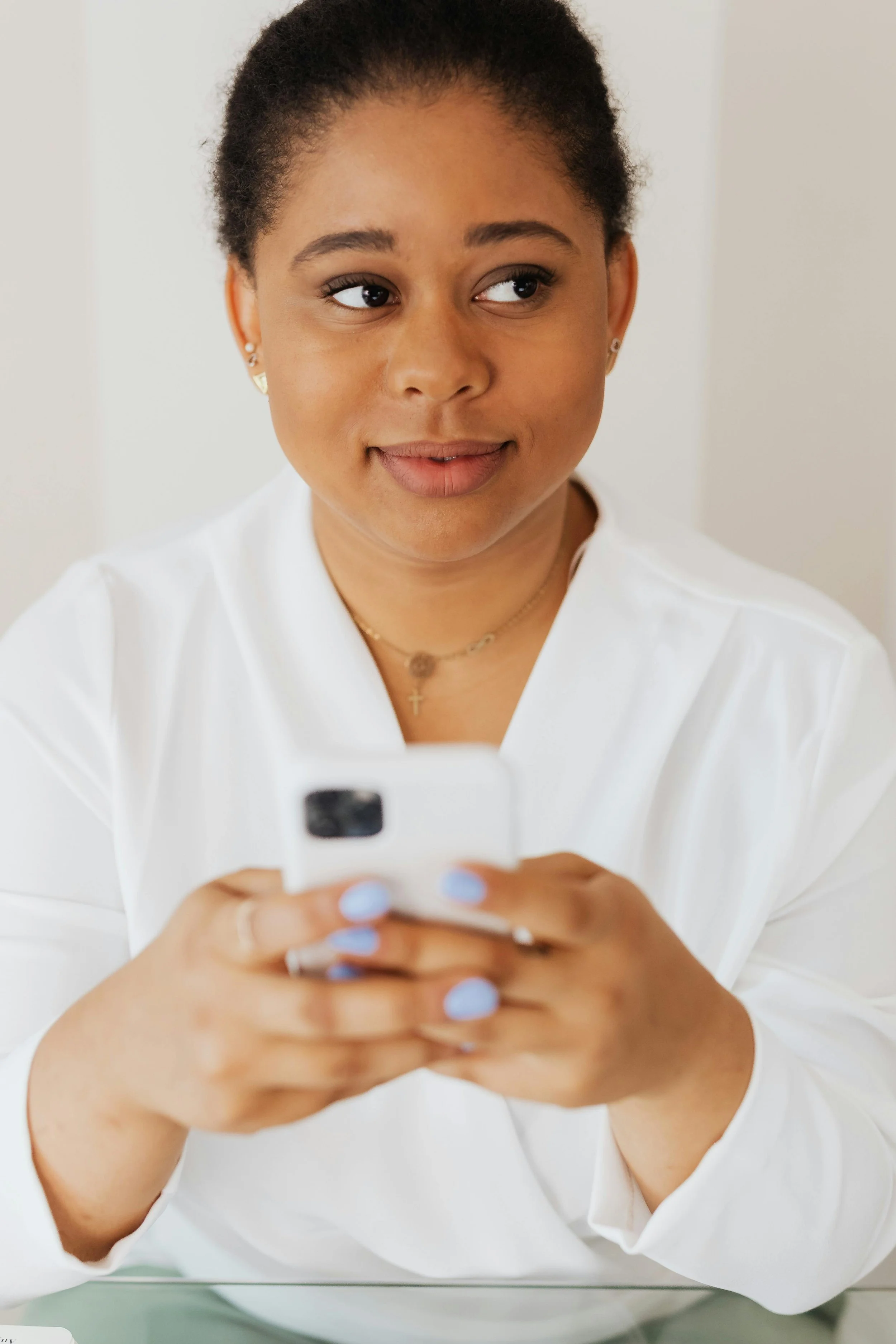 A woman with dark hair tied back, wearing a white blouse, looking at her phone with her nails painted light blue, and smiling.