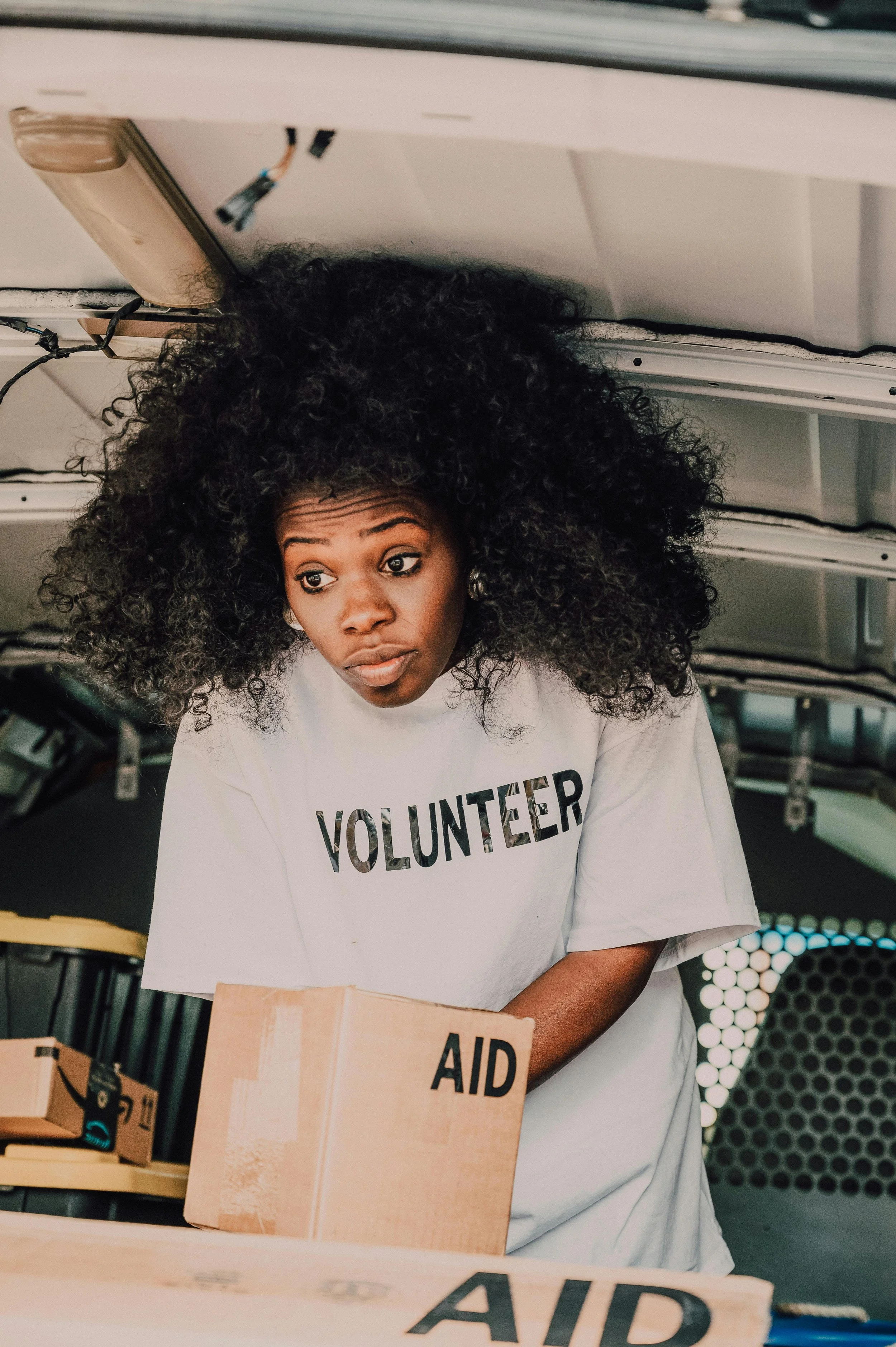 A woman with curly hair wearing a white t-shirt that says "VOLUNTEER" is organizing boxes labeled "AID" in what appears to be a storage or distribution center.
