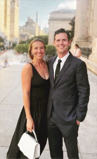 A smiling woman in a black dress holding a white handbag standing next to a smiling man in a suit, outdoors in an urban setting with historic architecture.
