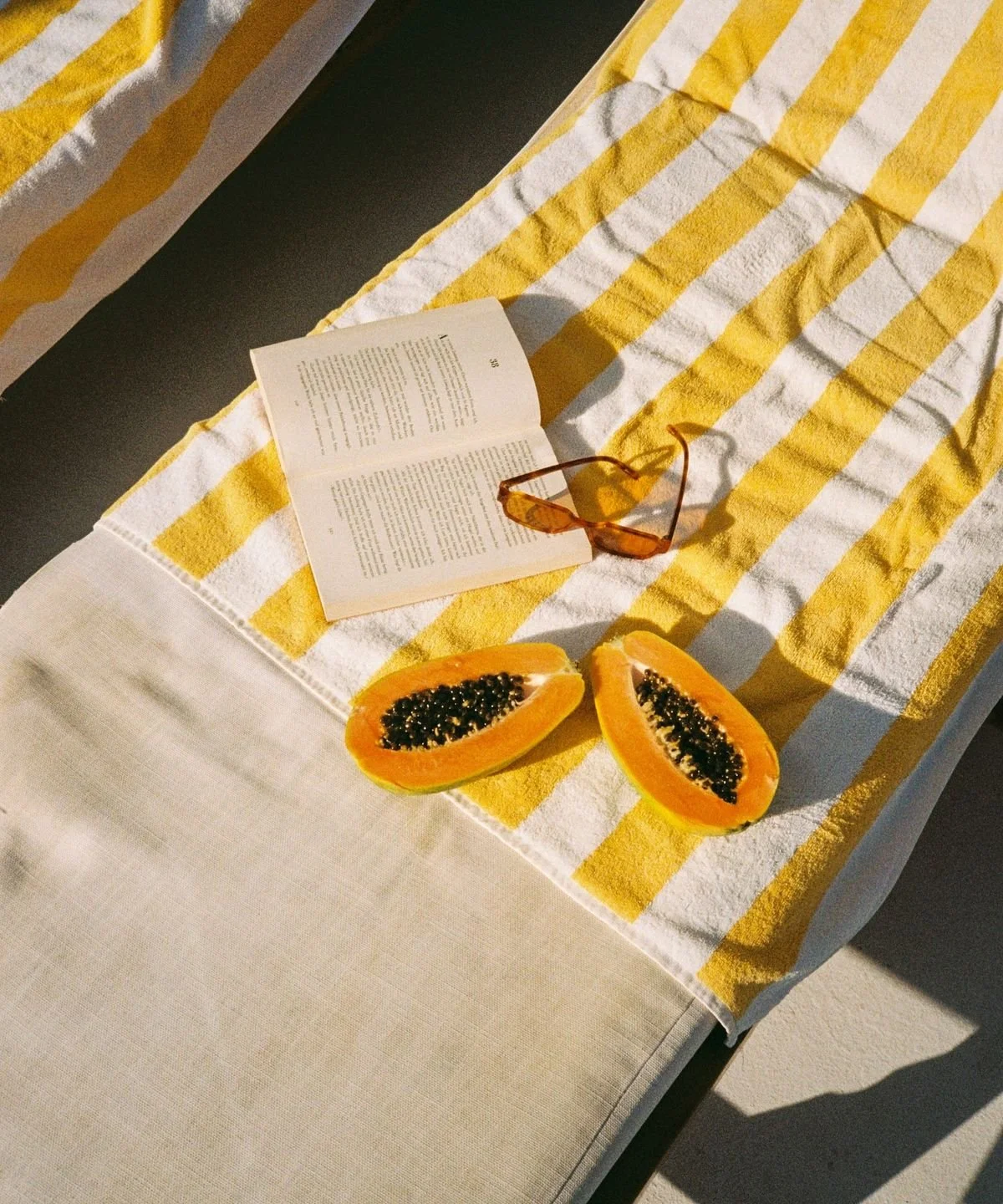 Une serviette de plage rayée de jaune et blanc posée sur une table, avec un livre ouvert, une paire de lunettes de soleil oranges, et deux moitiés de papaye. Santa Brevina