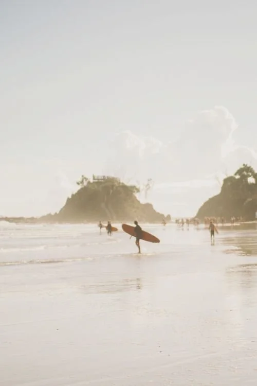 Sur la plage, plusieurs personnes dont un surfeur avec une planche de surf, et d'autres promenant le long du rivage, avec des îles ou rochers au loin et un ciel partiellement nuageux. Santa brevina