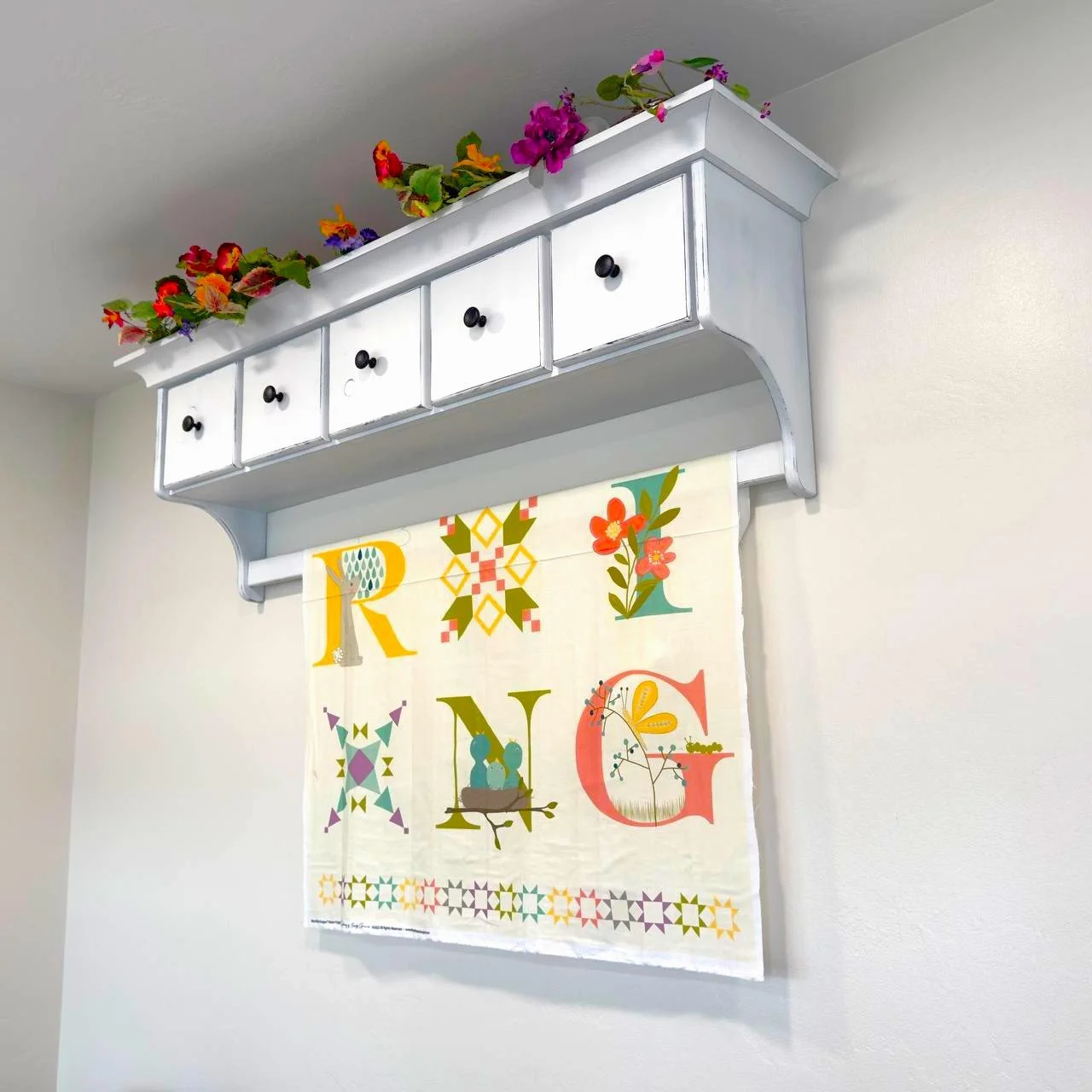 Wall shelf with small drawers and black knobs, decorated with pink, orange, purple, and yellow flowers on top, hanging above a colorful fabric wall hanging with the letters S, P, R, I, N, & G and various patterns and floral designs.