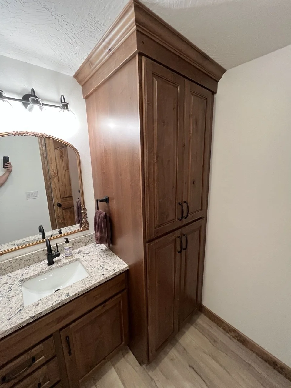 View of a bathroom vanity with a granite countertop, sink, faucet, mirror, and a tall wooden cabinet beside it.