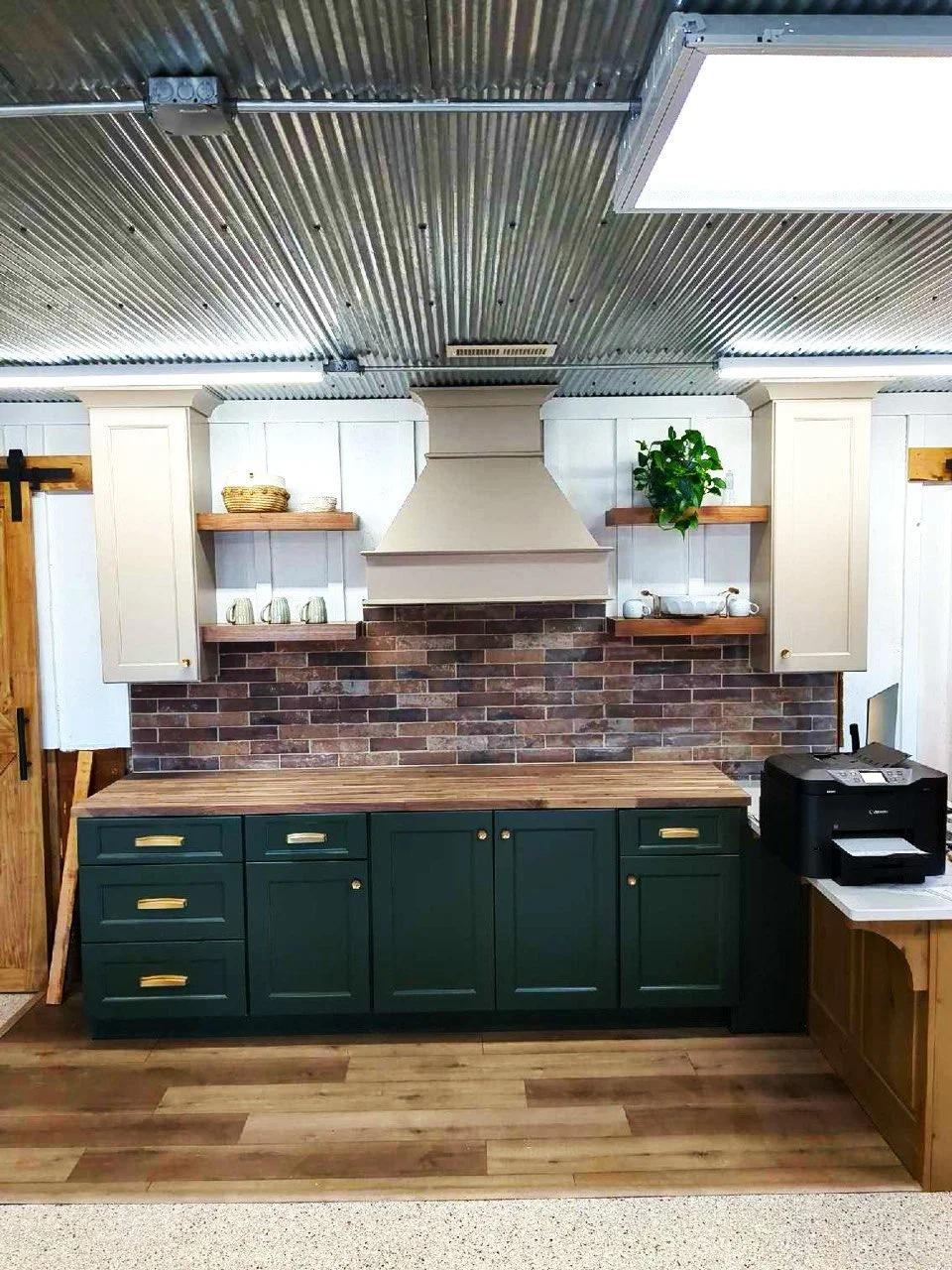 Kitchen with dark green cabinets, wooden countertops, a brick backsplash, white upper walls, open shelves with decorative items, a white range hood, potted plant, and a black printer on a small table.