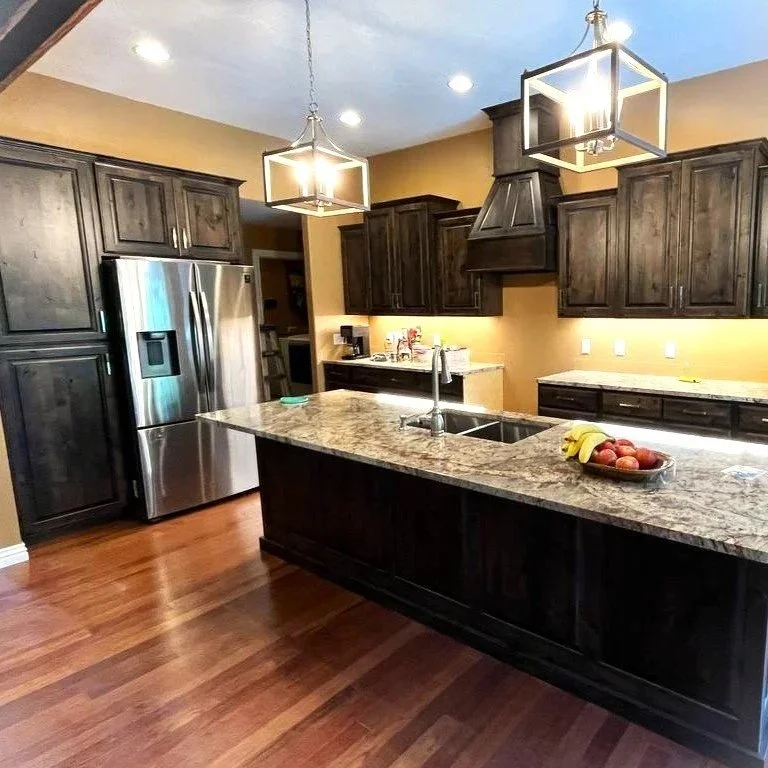 Modern kitchen with dark wood cabinets, stainless steel refrigerator, granite countertop island, hardwood floors, and contemporary pendant lighting.