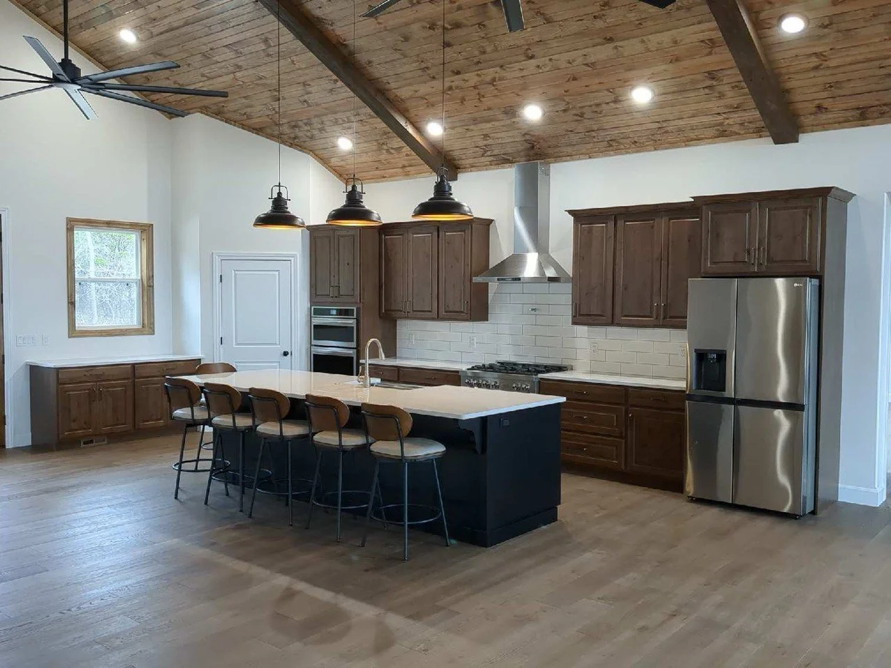 Modern kitchen with wooden cabinets, stainless steel refrigerator, white subway tile backsplash, and an island with a white countertop and five chairs. Wooden vaulted ceiling with recessed lighting and black pendant lights.