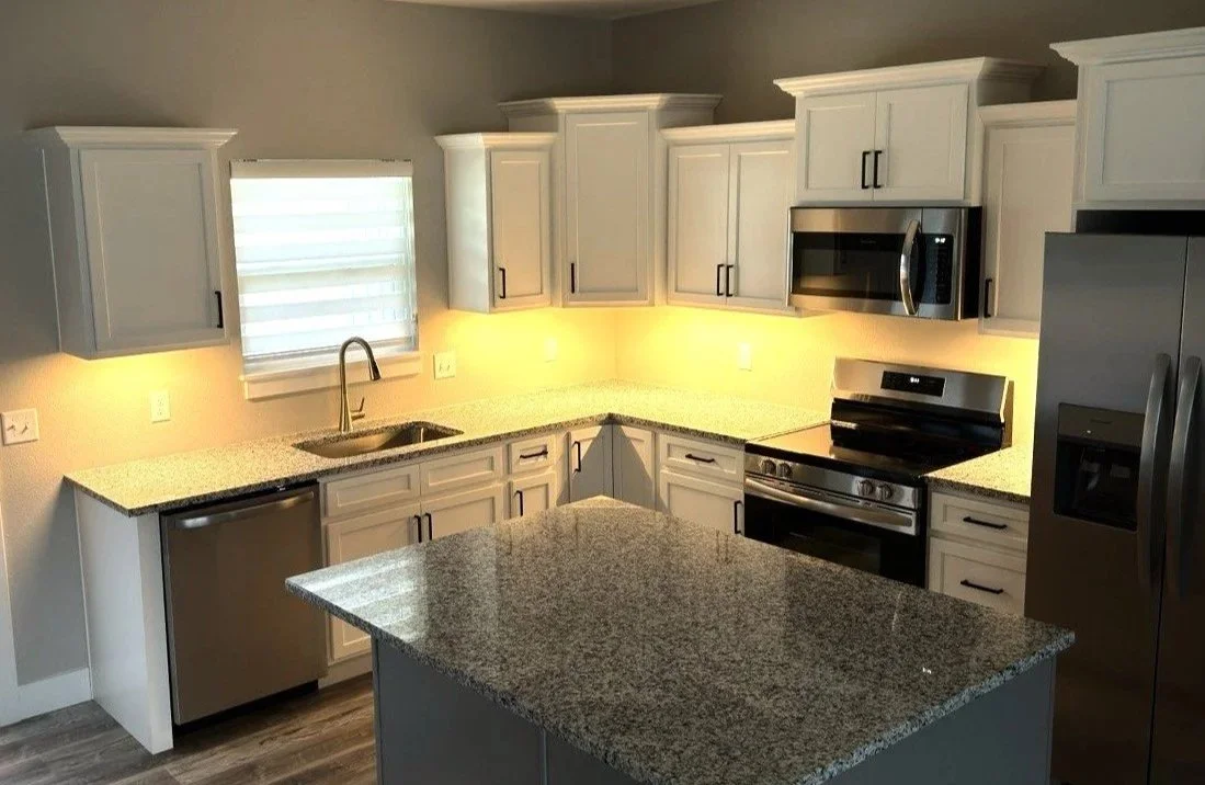 Modern kitchen with gray cabinets, granite countertops, stainless steel appliances including refrigerator, microwave, oven, and dishwasher, with a small kitchen island in the foreground and a window above the sink.