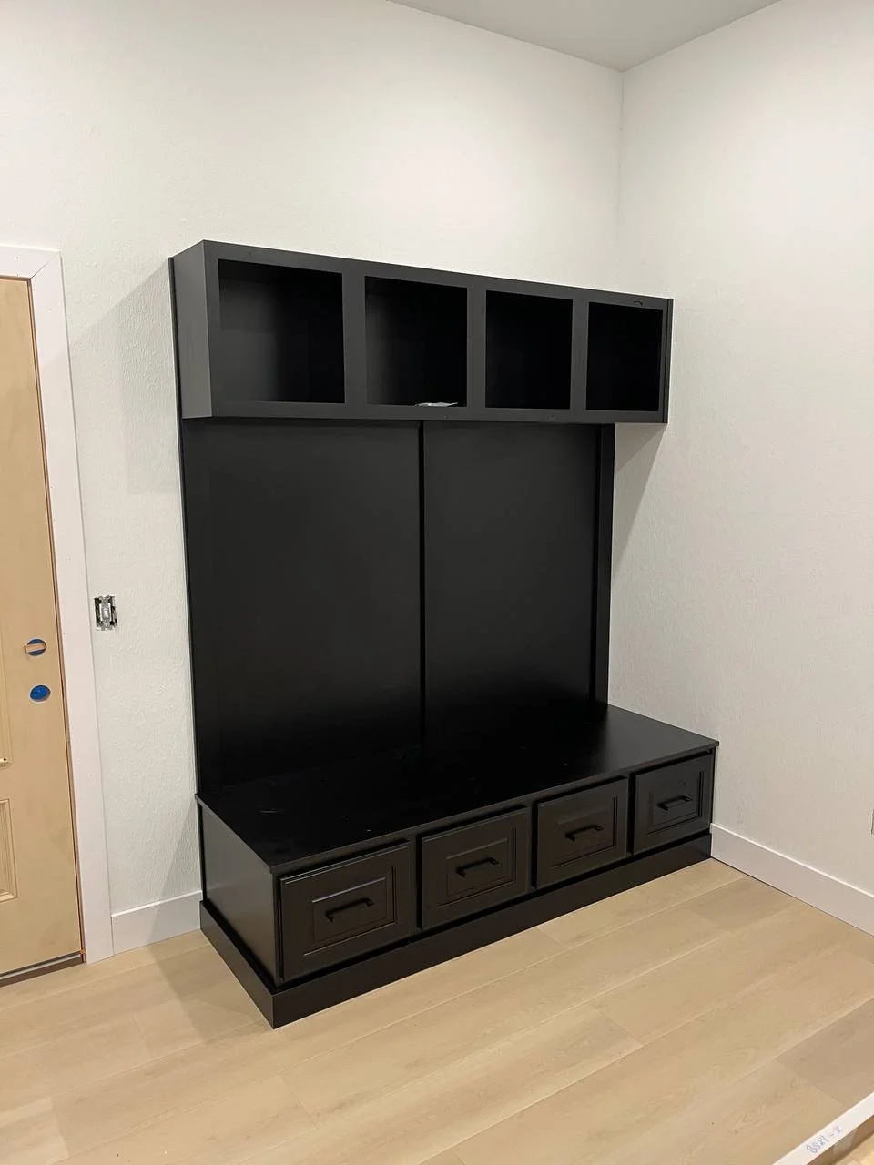 Black wooden storage bench with four drawers at the bottom, a large back panel, and open shelves above, positioned against a white wall in a room with light wood flooring.