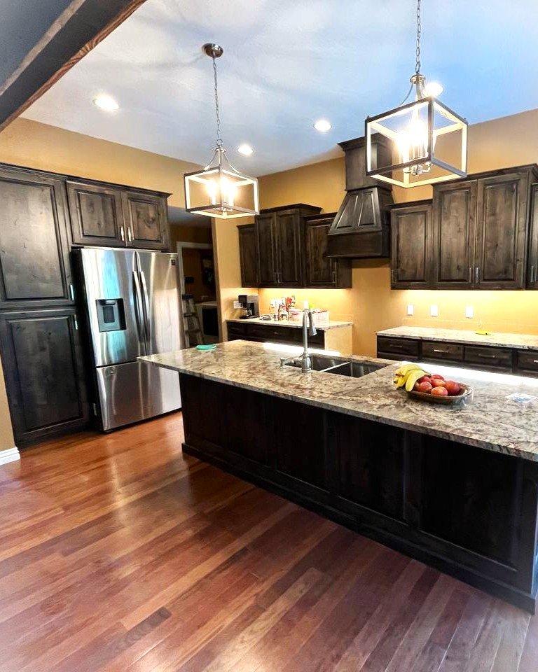 Modern kitchen with dark wood cabinets, granite countertops, stainless steel refrigerator, island with sink, wooden flooring, and contemporary pendant lights.