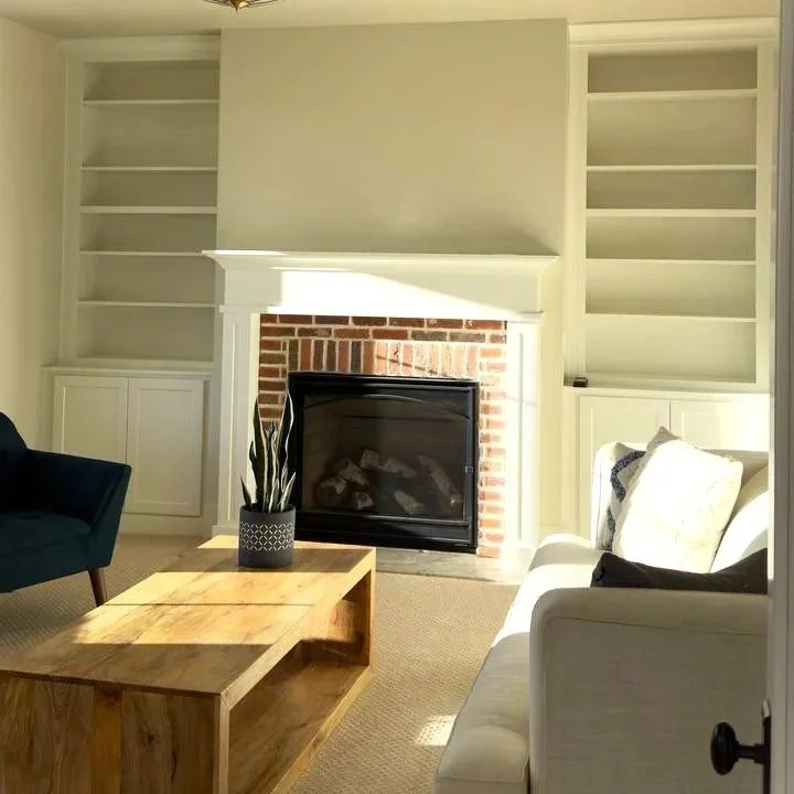 Living room with fireplace, built-in shelves, a white couch with pillows, a dark armchair, a wooden coffee table with a potted plant, and a brick fireplace with a black frame.
