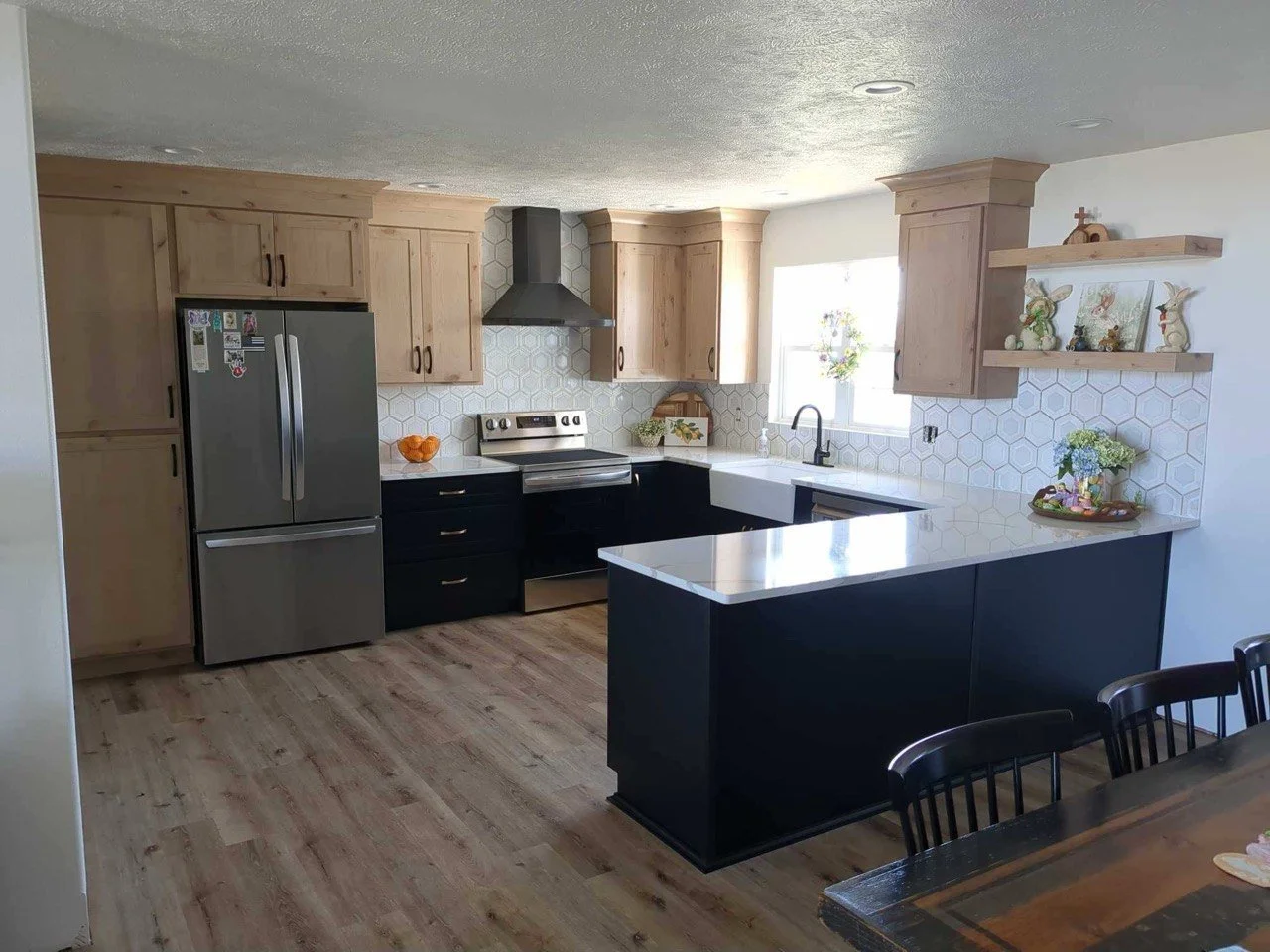 Modern kitchen with light wood cabinets, black lower cabinets, a stainless steel refrigerator, a white farmhouse sink, and a white quartz countertop island. Decor includes bunny figurines, potted plants, and a hanging flower wreath by the window. Hexagonal tile backsplash and wood flooring complete the look.