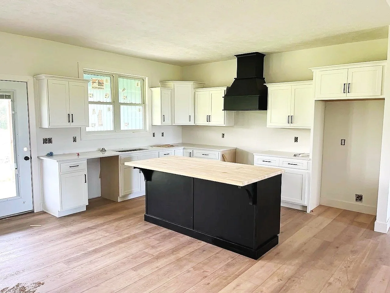 Empty white kitchen with black island, white cabinets, black range hood, hardwood floors, and a window above the sink.