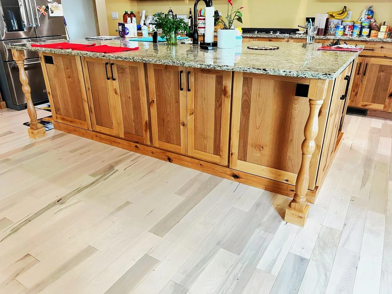 Kitchen island with wooden cabinets and granite countertop, with various kitchen items and plants on top, in a bright kitchen.