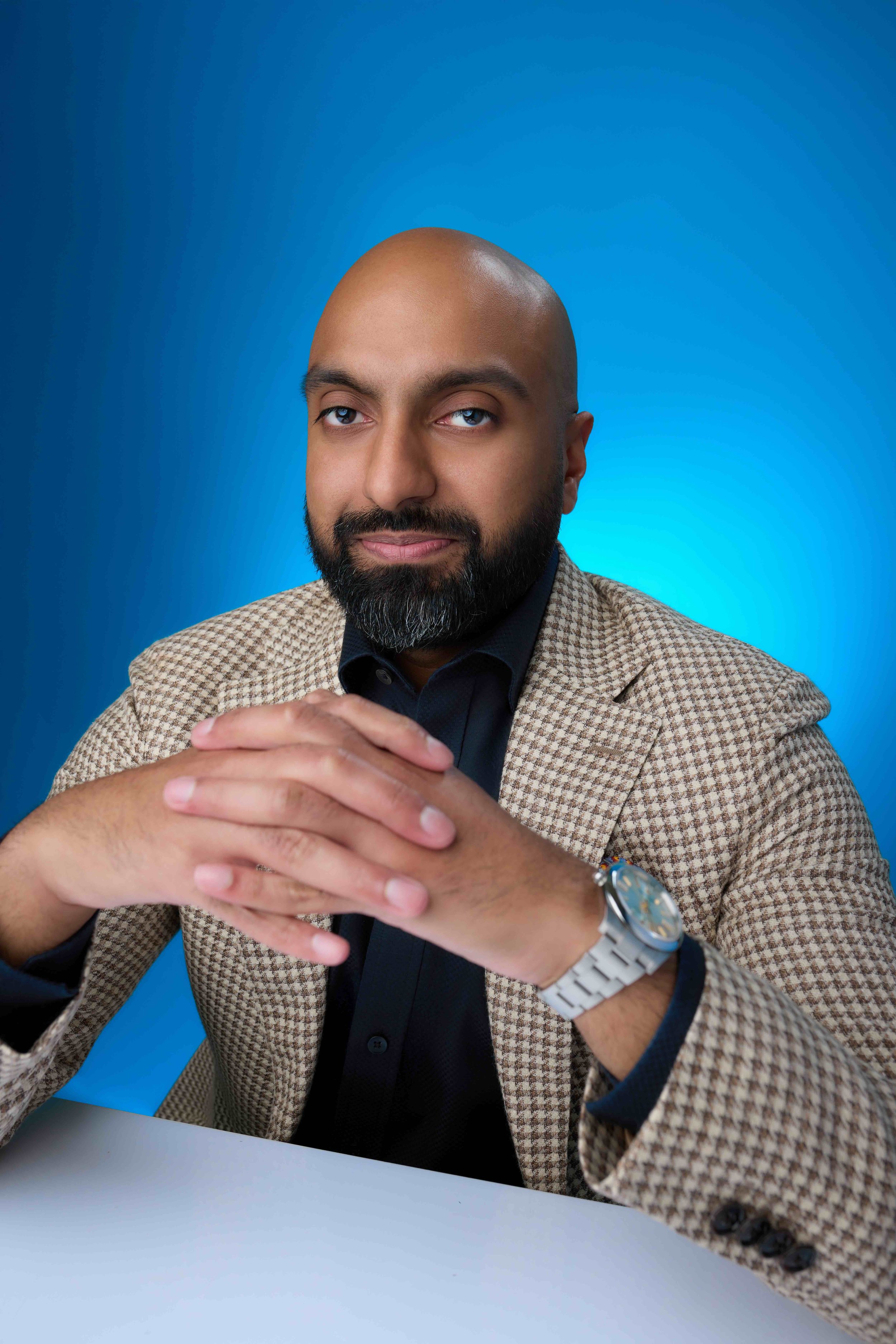 A man with a bald head and a beard, wearing a beige checkered blazer, black shirt, and a watch, sitting at a white table with a bright blue background.