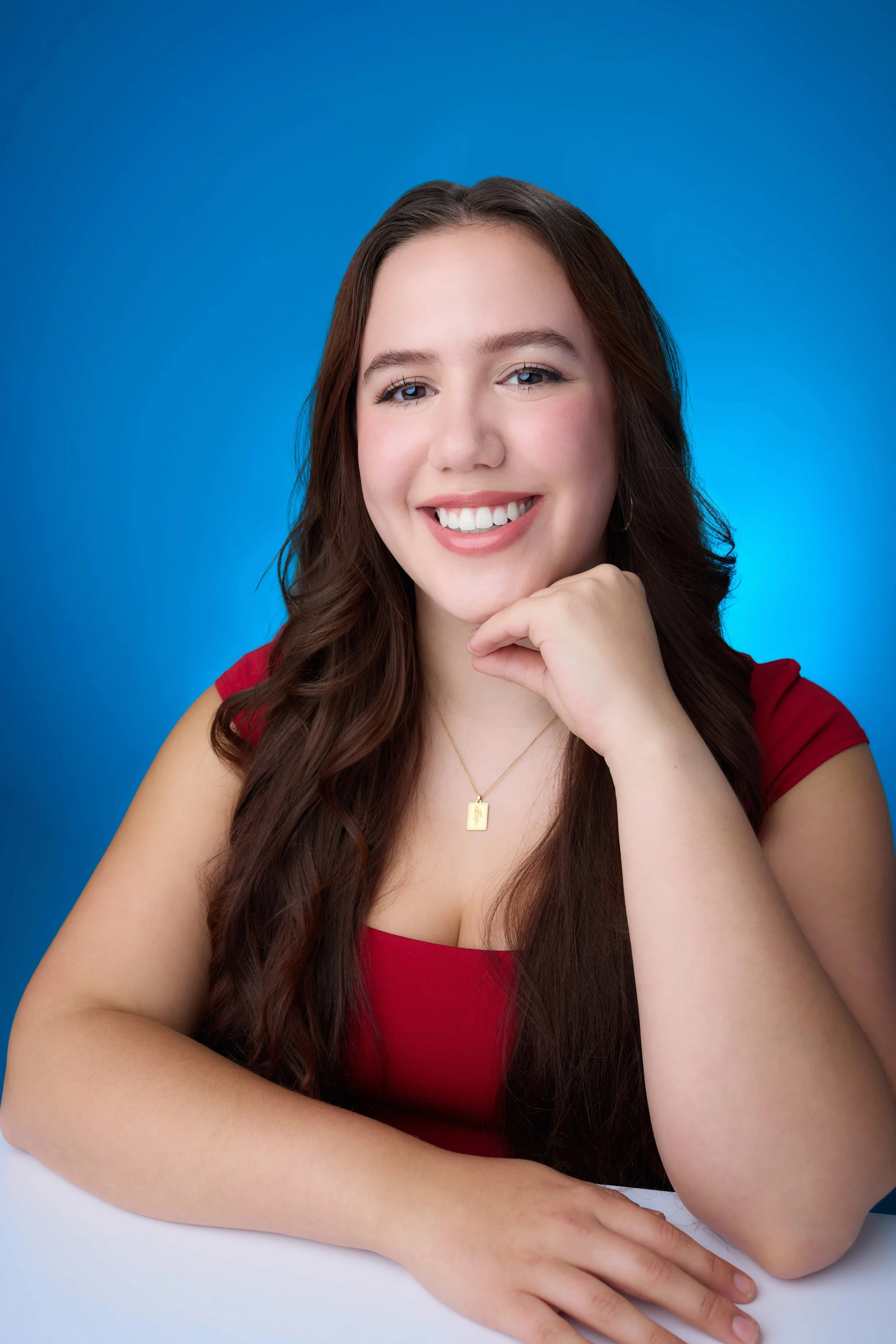 Portrait of a young woman with long brown hair and blue eyes, smiling and resting her chin on her hand, wearing a red top and gold necklace, against a blue background.