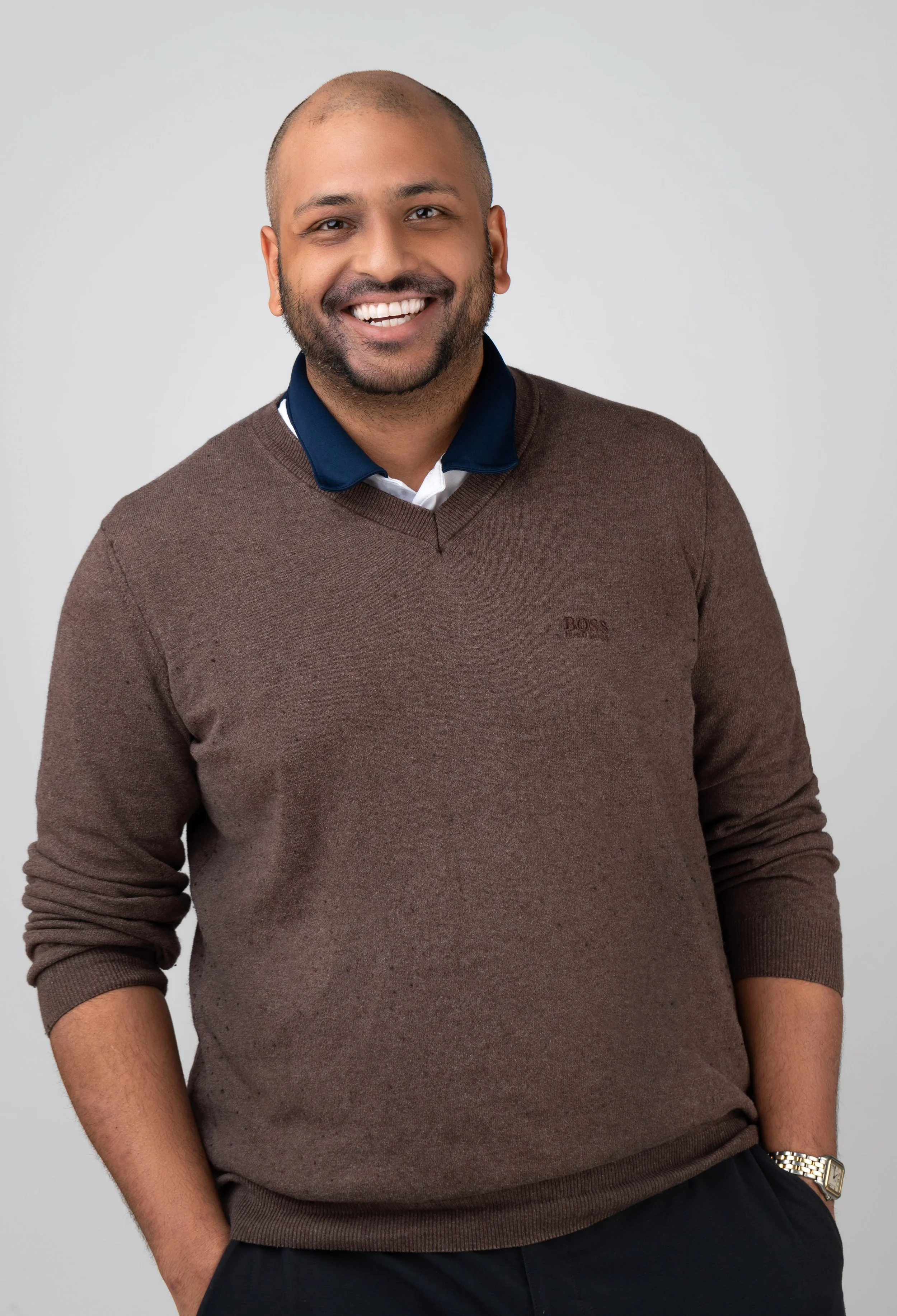 A smiling man with a beard and shaved head wearing a brown sweater over a dark collared shirt, standing against a plain light gray background.