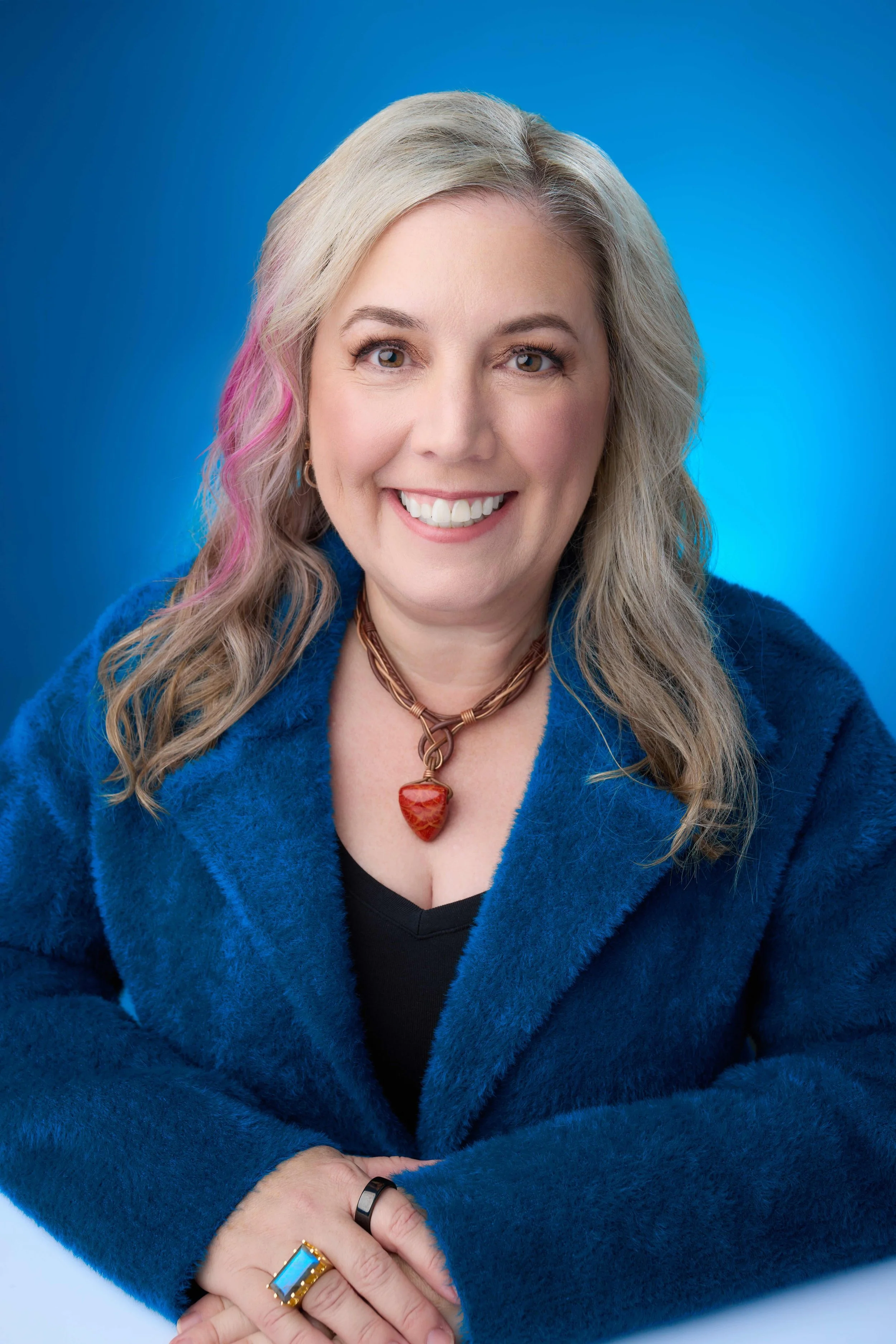 A woman with blonde hair and a pink streak in her hair smiling, wearing a blue furry coat, a black top, a necklace with a red heart-shaped pendant, a black ring, and a gold ring with a blue stone, with her hands clasped in front of her against a blue background.
