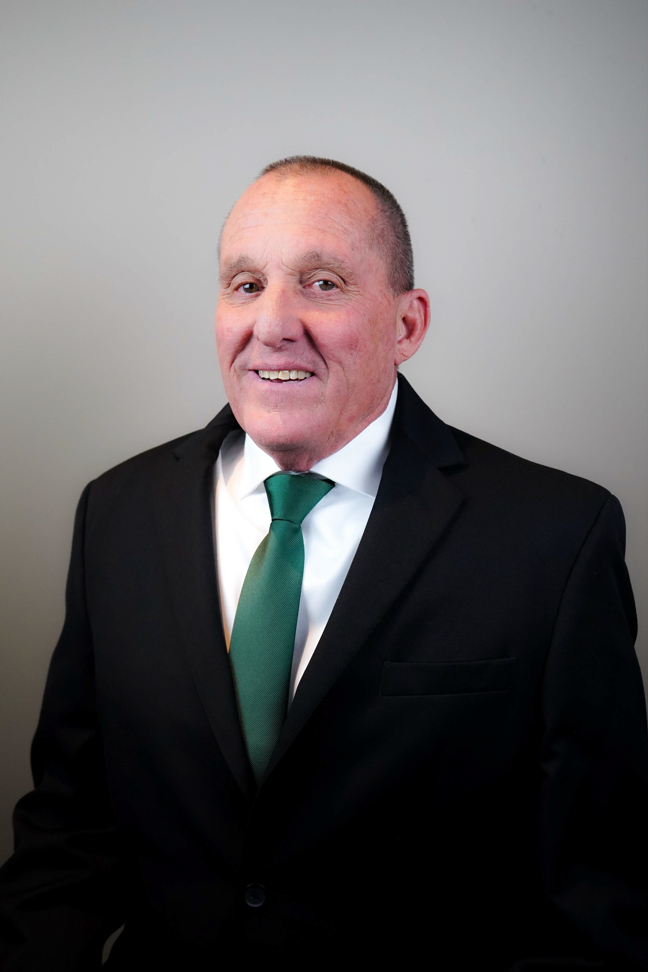 A middle-aged man wearing a black suit, white shirt, and green tie, smiling with a neutral gray background.