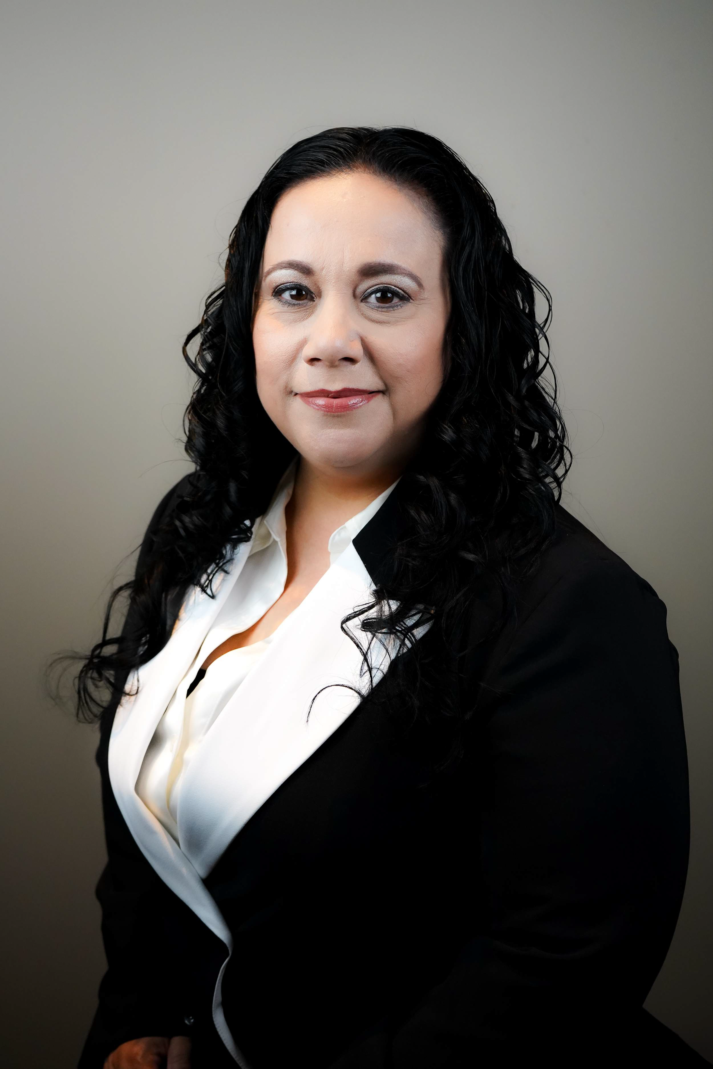 A woman with long black curly hair wearing a black blazer and white blouse standing against a plain gray background.