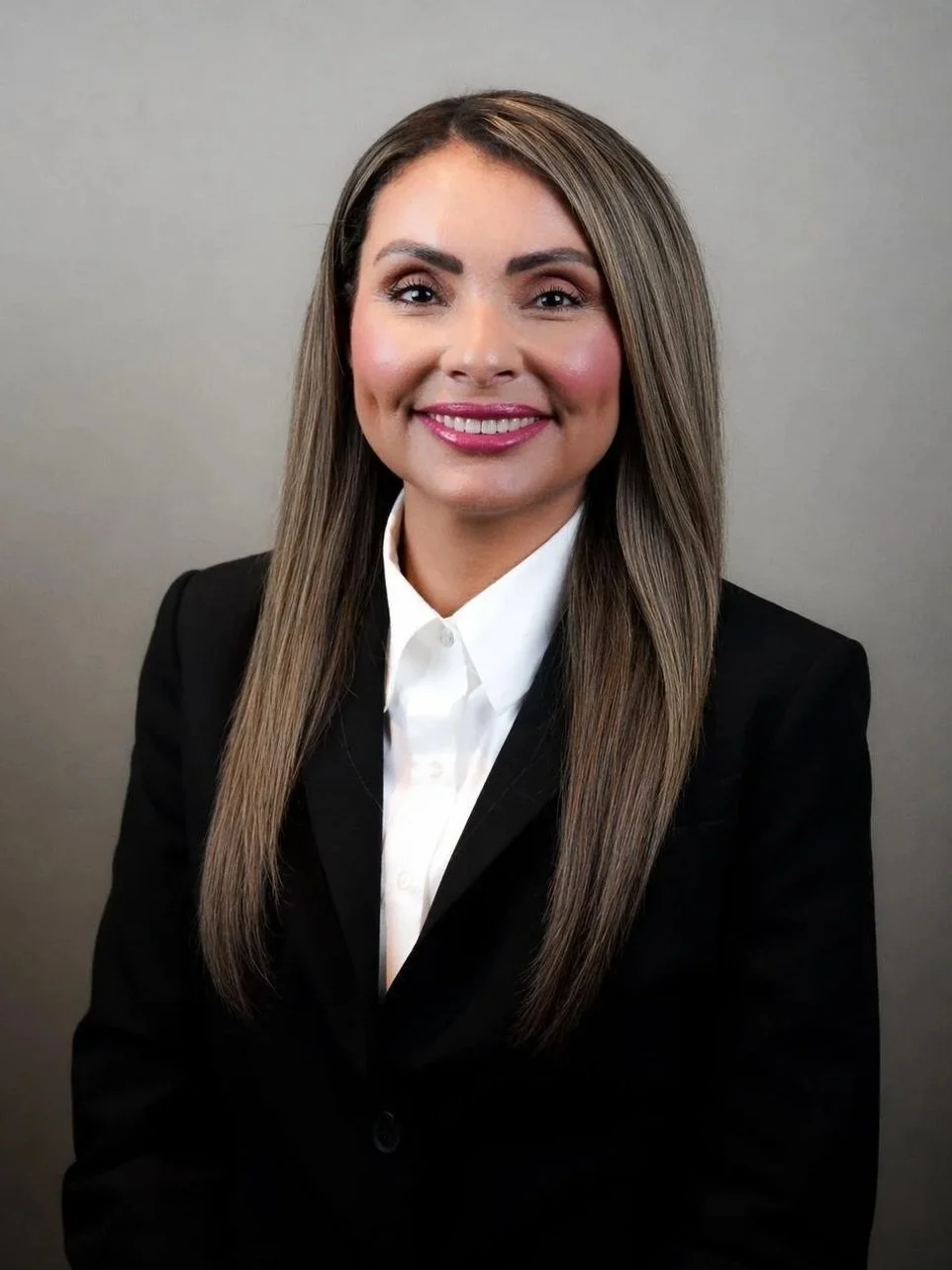 Professional woman with long straight hair wearing a black blazer and white shirt, smiling at the camera against a plain gray background.