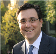 A smiling man in a suit outdoors with green trees in the background.