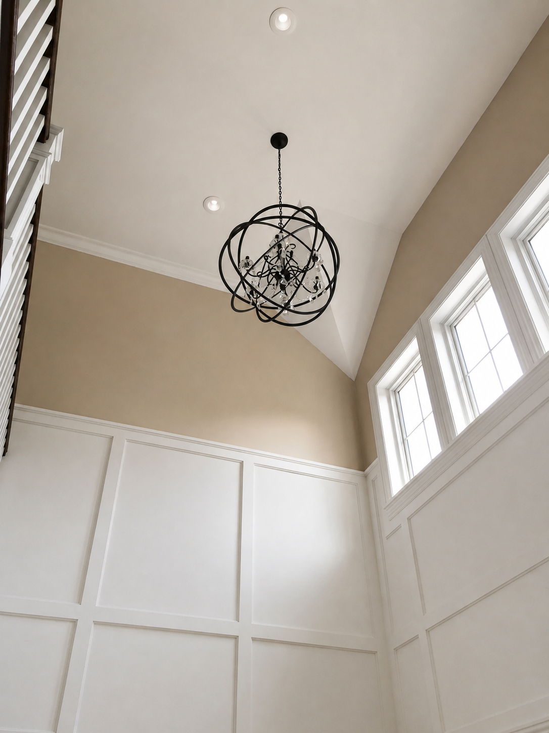 Interior ceiling view featuring a modern chandelier hanging from a black chain, recessed ceiling lights, and large windows on the right letting in natural light. The walls are beige with white wainscoting and crown molding.