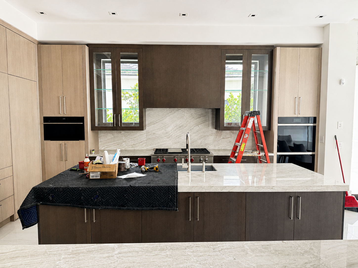 Modern kitchen with a large marble island, dark and light wooden cabinets, glass-front display cabinets, stainless steel oven, and a red step ladder. Construction tools and supplies are on the island, indicating ongoing work.