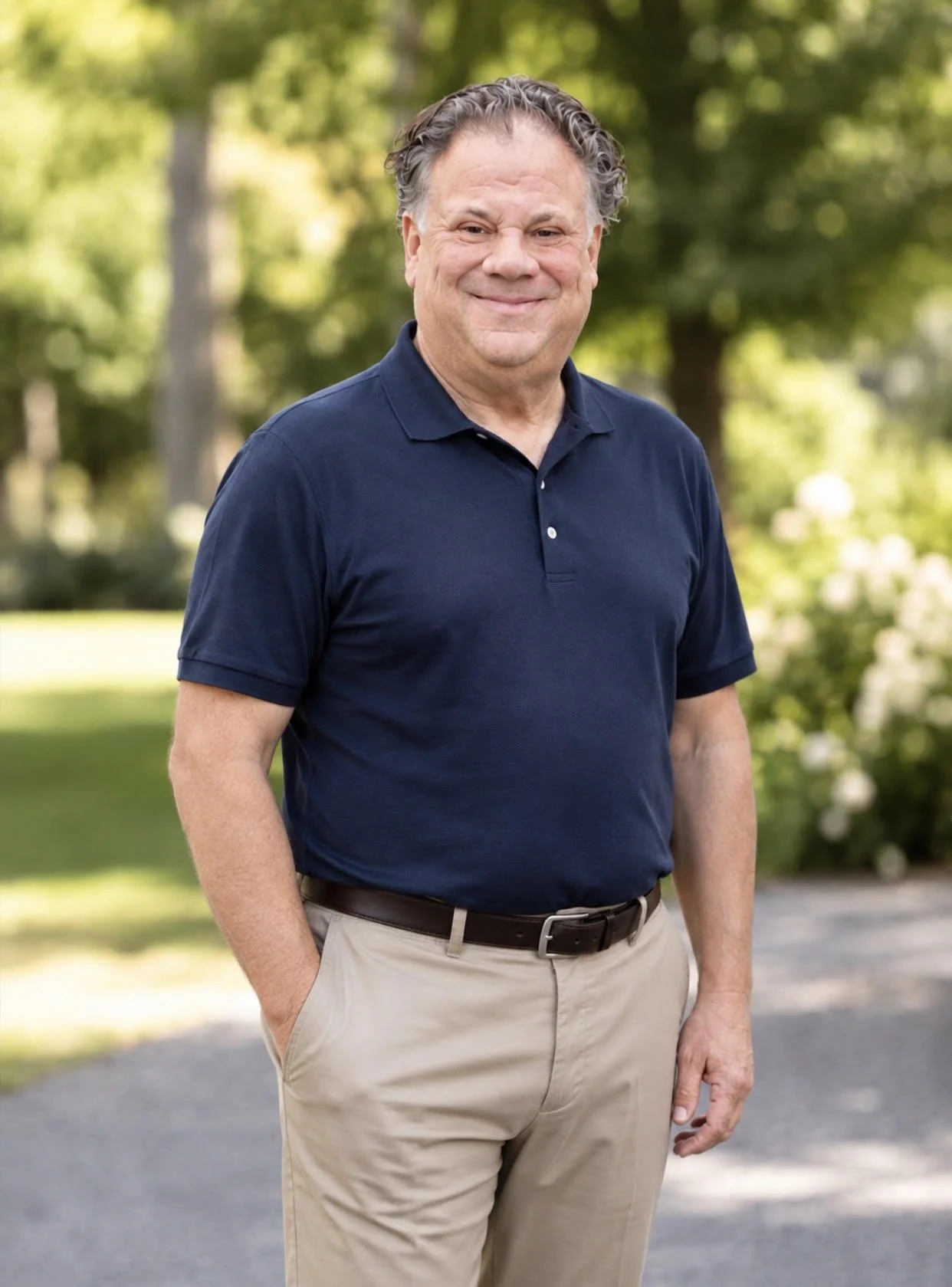 A middle-aged man with wavy hair smiling and standing outdoors in a park, wearing a navy blue polo shirt and khaki pants.