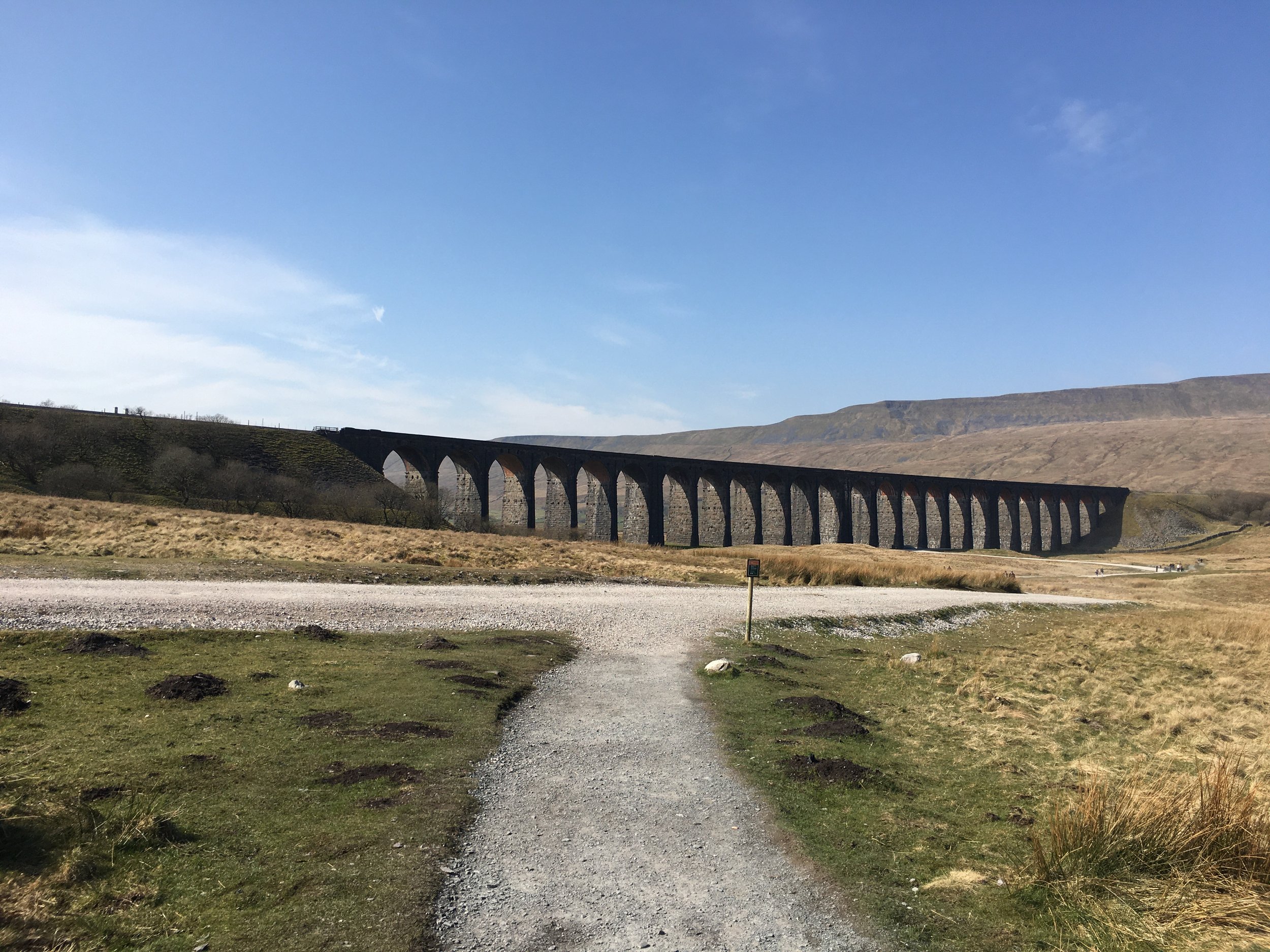 A landscape view of a mountainous area with a stone railway viaduct with multiple arches crossing a valley, under a bright blue sky with some clouds.