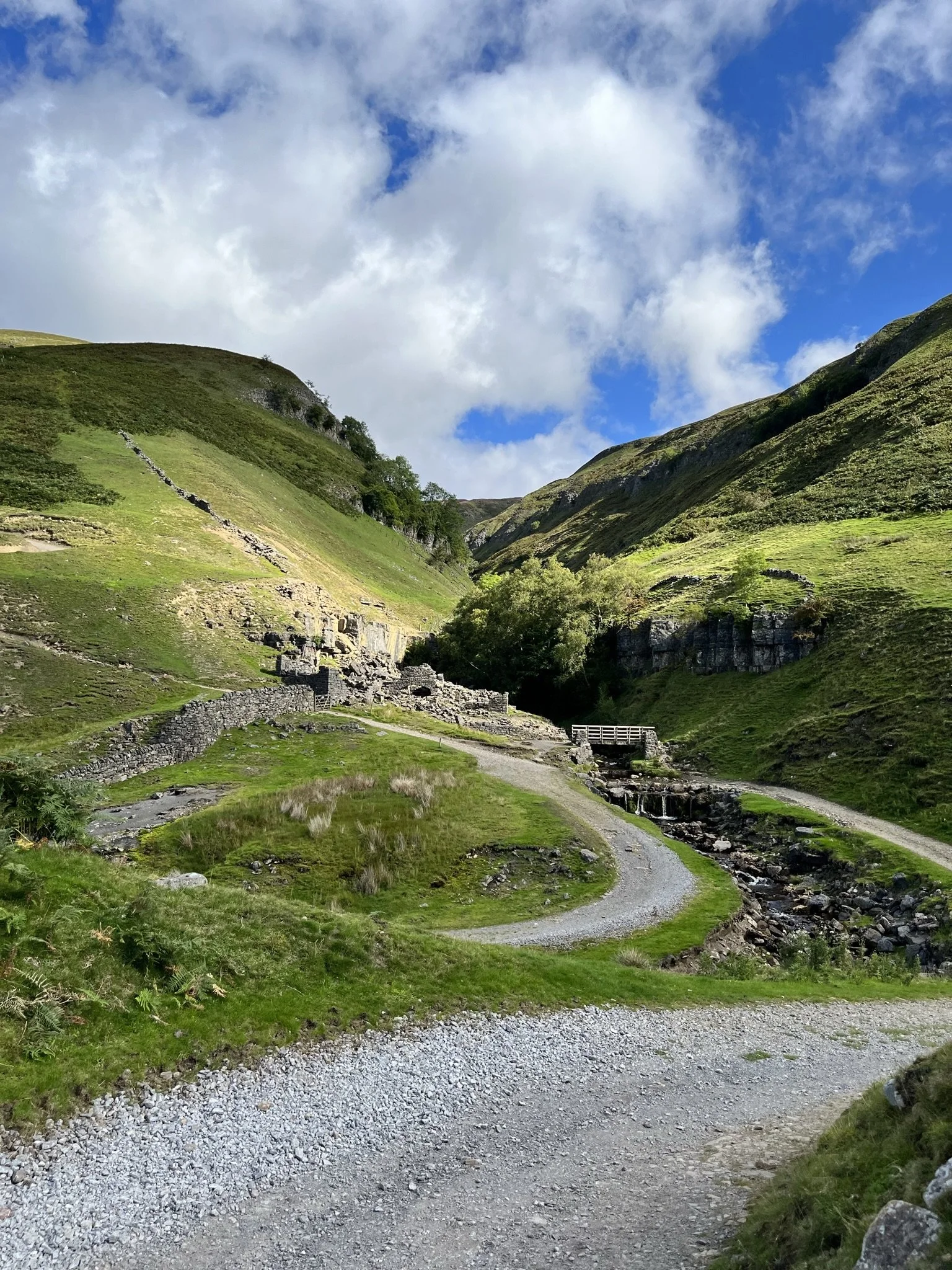 A scenic mountain landscape with green grassy slopes, rocky terraces, and a dirt path winding along a river or stream, under a partly cloudy sky.