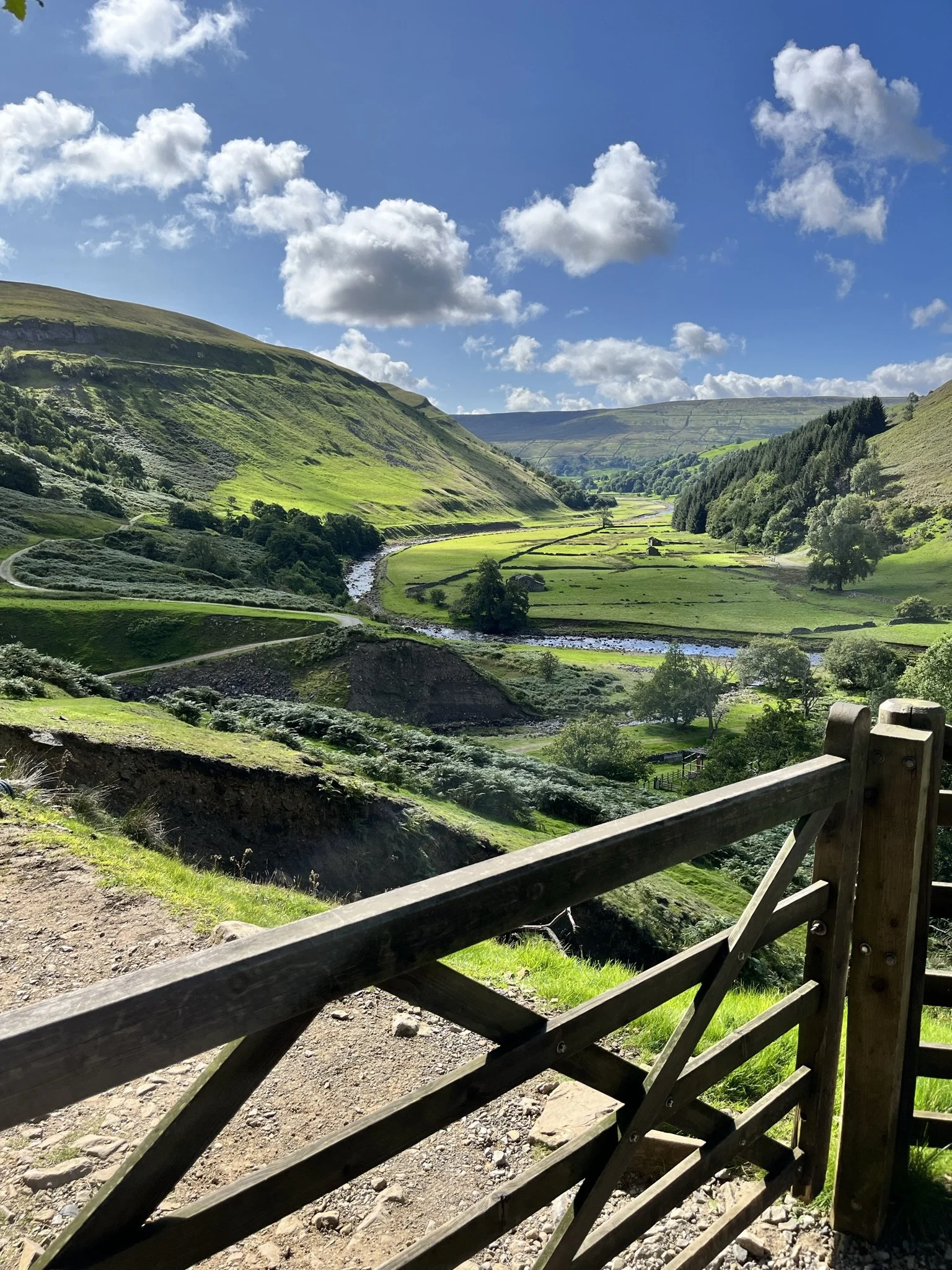 A scenic view of a lush green valley with rolling hills, a winding river, and a few structures, viewed from a wooden fence on a sunny day with a partly cloudy sky.