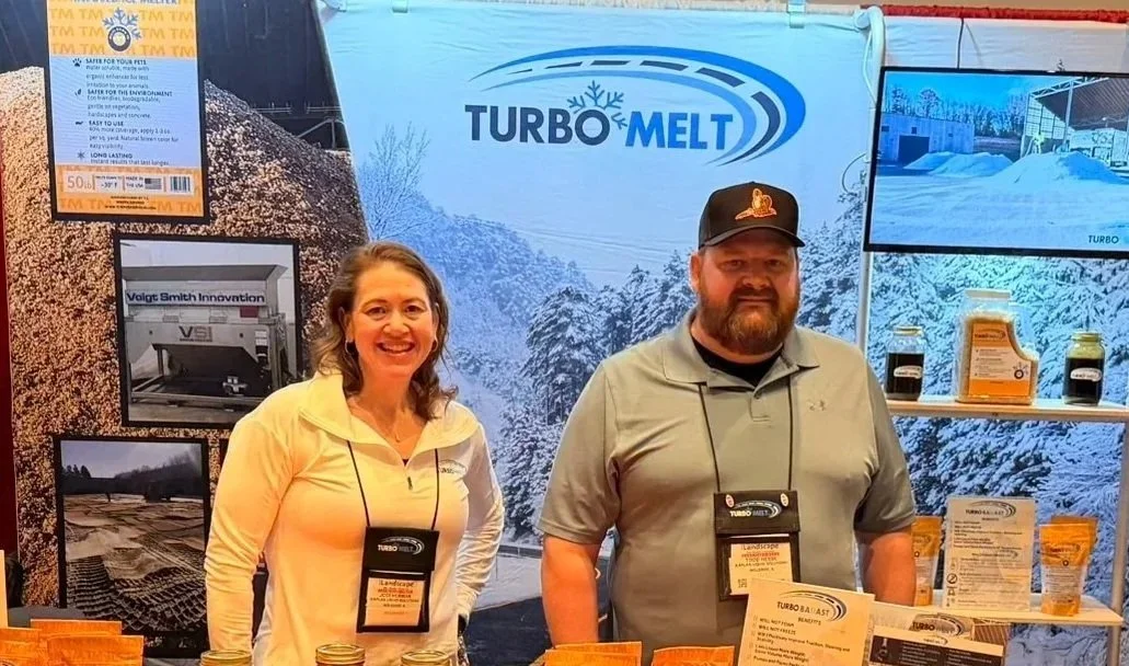 Two people standing in front of a winter-themed trade show display for Turbo Melt, with products on shelves and promotional material behind them.