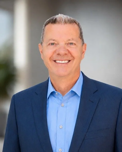 A business man with short hair, wearing a blue dress shirt and a dark blue blazer, smiling outdoors against a blurred background.