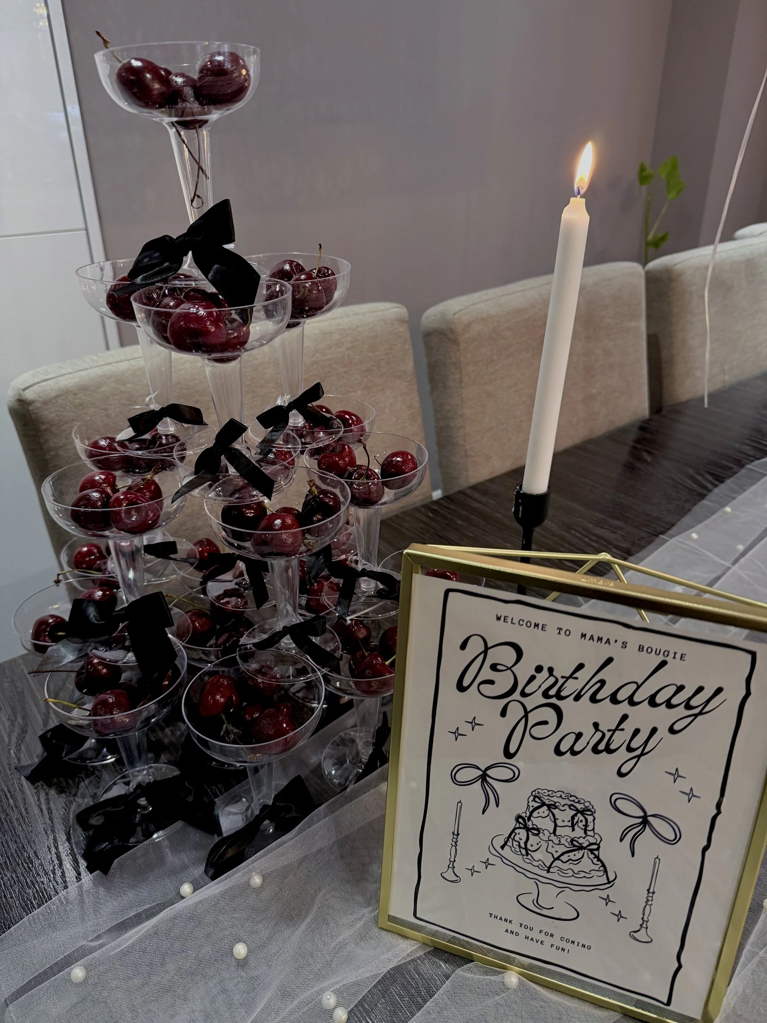 A birthday party table with a sign, decorated with cherries in clear cups, and a white candle in a candle holder, with beige chairs and a gray wall in the background.