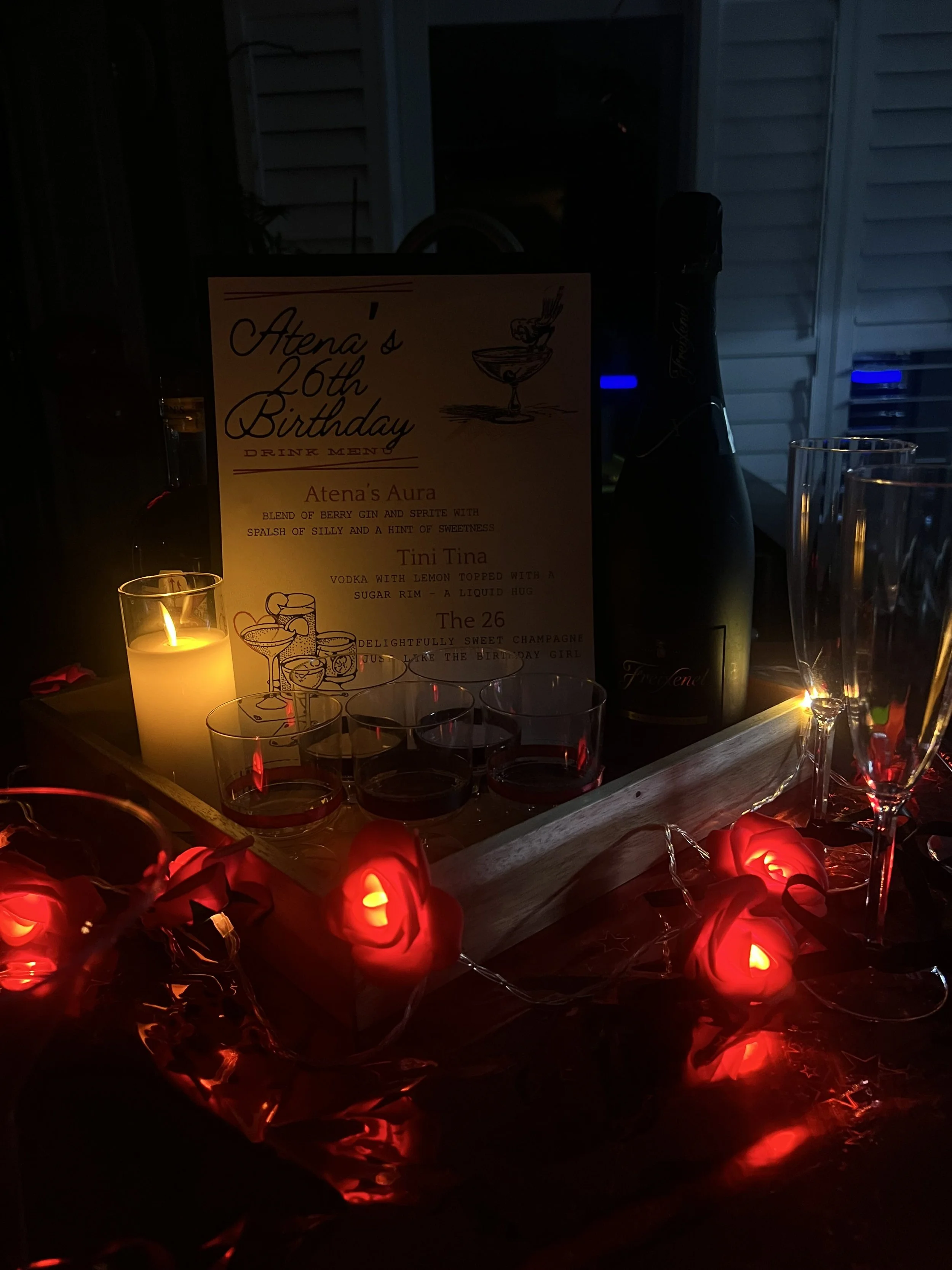 A dimly lit outdoor table decorated for a birthday celebration with a sign, candles, glasses, a bottle of champagne, and red rose-shaped lights.